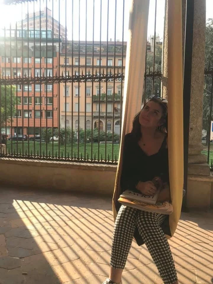 A young woman sitting on a step outdoors, partially hidden behind a curtain, holding a book, with sunlight casting shadows through a metal fence, with buildings in the background.