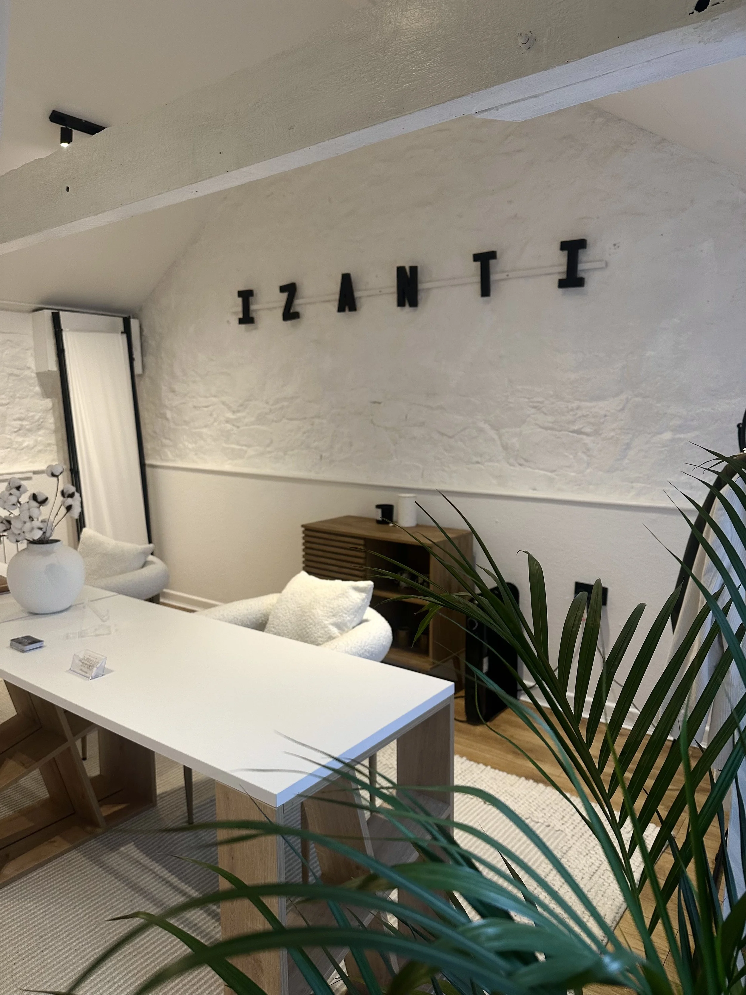 Modern office space with a white table, white chairs with plush cushions, a decorative white vase with flowers on the table, and a textured white wall with black letters spelling 'IZANTI' hanging above. There are plants with long green leaves in the foreground, and a small wooden shelf with a roll of paper and other items in the background.
