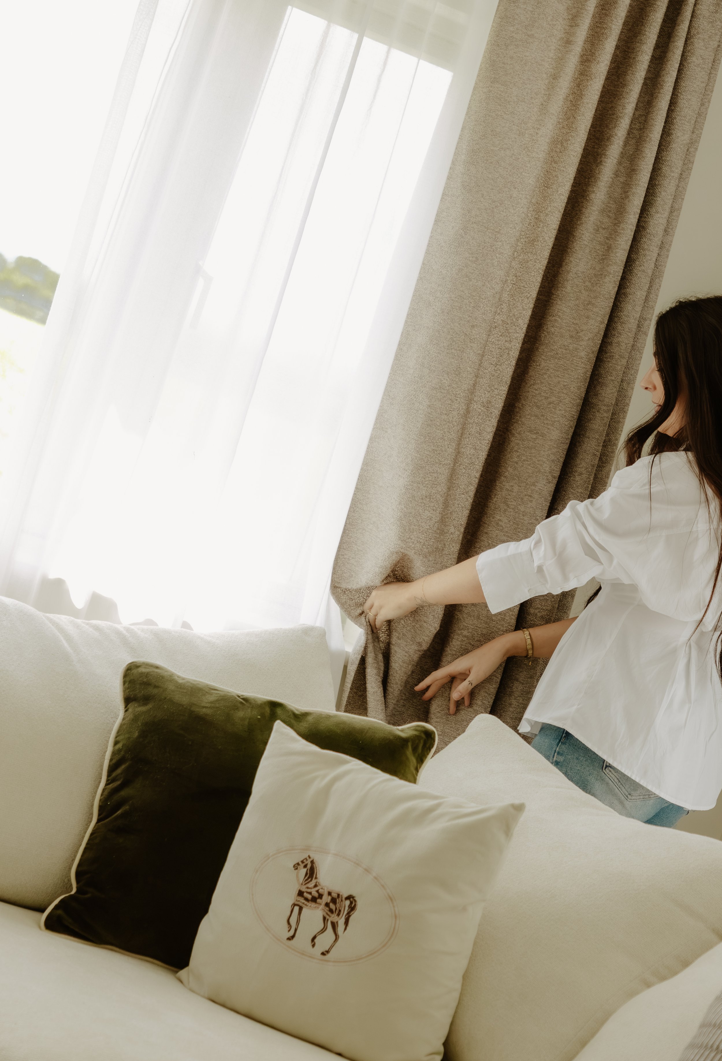 A woman in a white shirt adjusts beige curtains near a large window with white sheer curtains. In the foreground, there are pillows on a cream-colored couch, including one with a horse design.