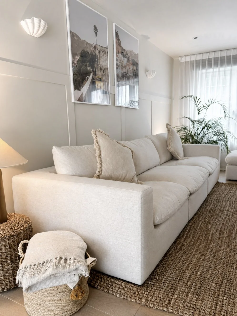 A living room with a cream-colored sofa, matching pillows, a woven basket with a blanket, a potted plant near a window, and framed seaside landscape photographs on the wall.