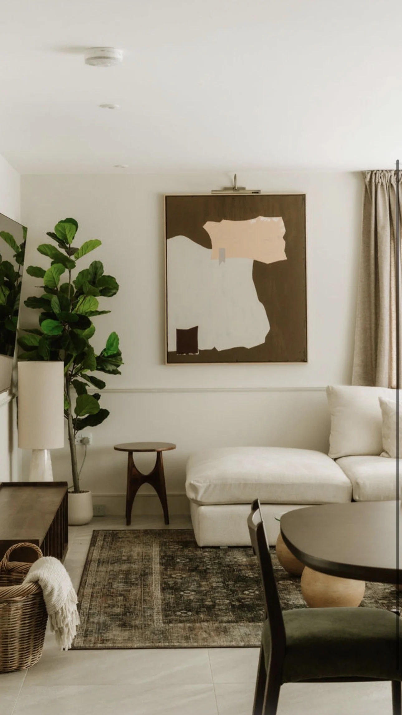 A living room with a large abstract painting hanging on a white wall, a potted plant on the left, a small round wooden side table, a white sofa with cushions on the right, a dark rug on the floor, a wooden table with a rounded edge in the foreground, and beige curtains on the window.