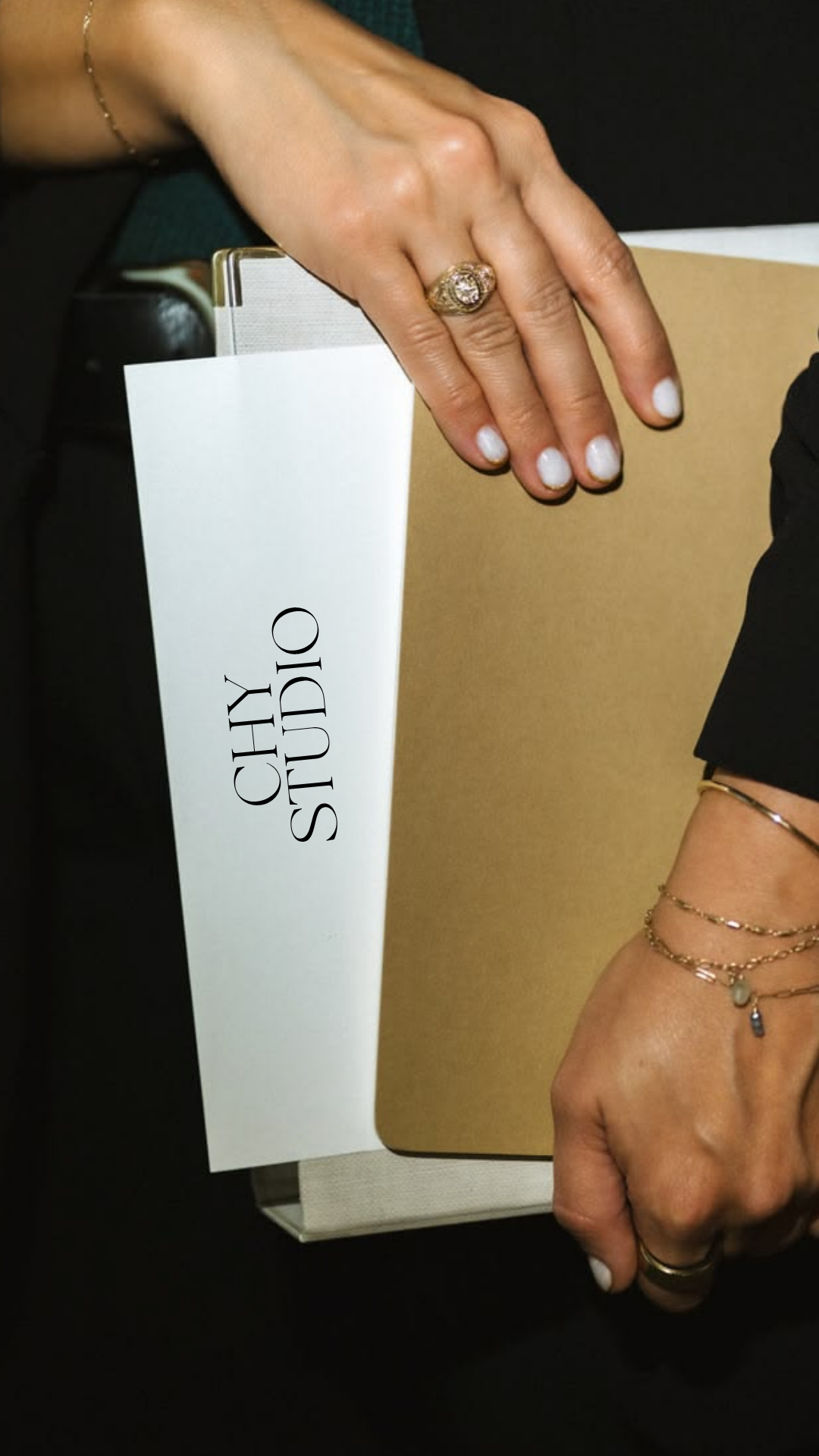 Close-up of a woman wearing multiple rings and bracelets, holding a Chloé Studio shopping bag, with a beige notebook and a black surface in the background.