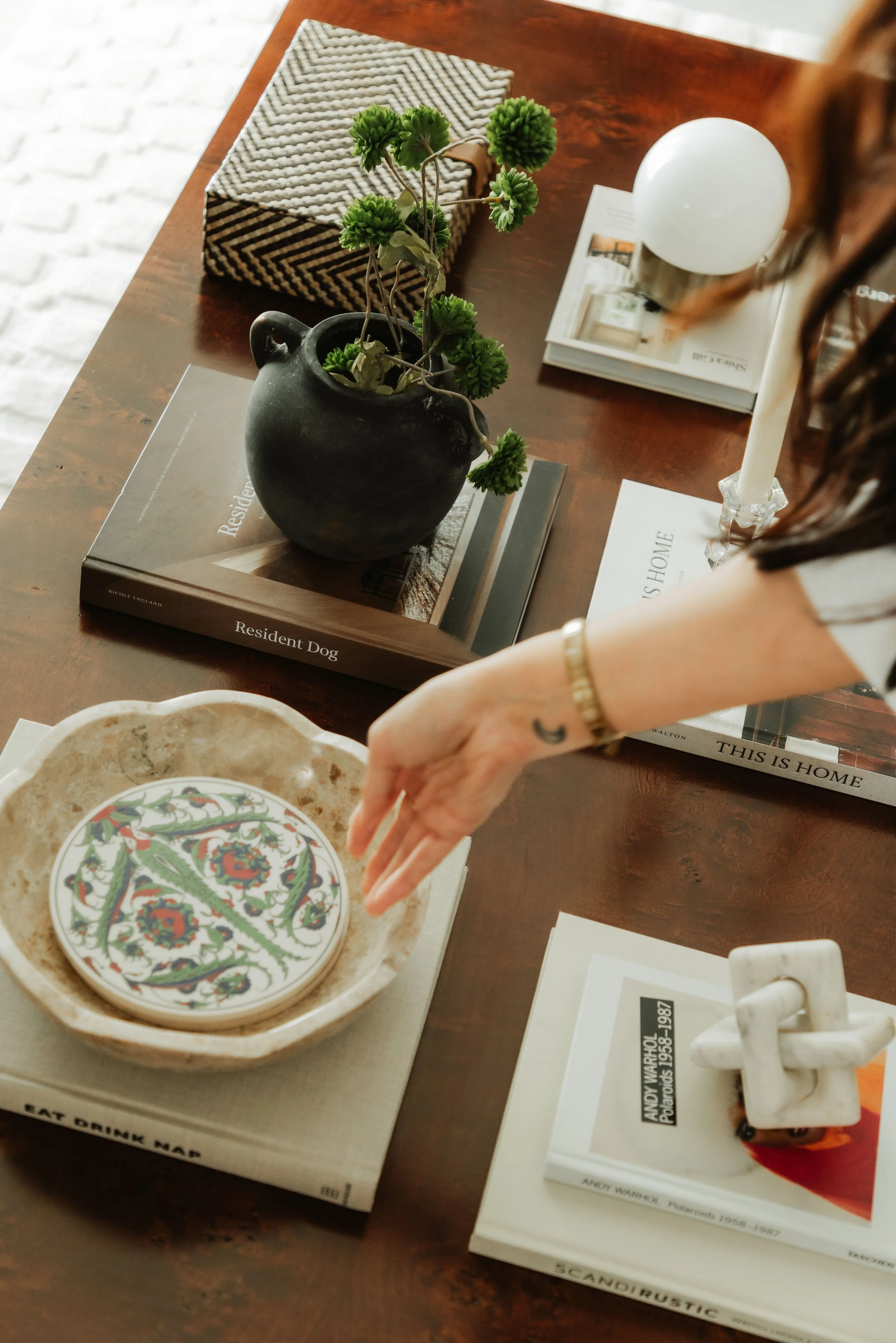 A wooden table with a black ceramic vase holding a small green plant, a patterned tissue box, a white candle, a magazine, and a decorative dish with colorful designs.