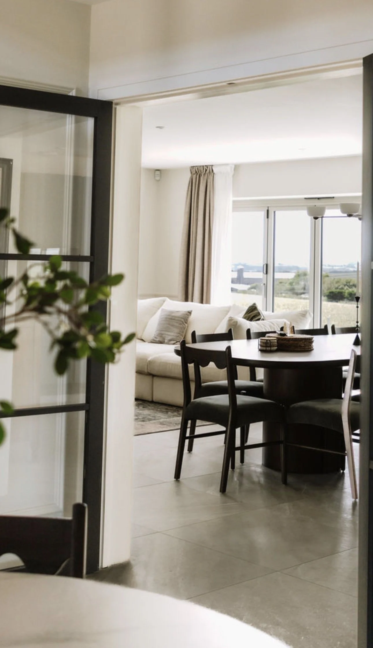 View of a dining area with a black table and chairs and a living room with a beige sofa, pillows, large glass sliding doors, and curtains, seen through an open sliding door.