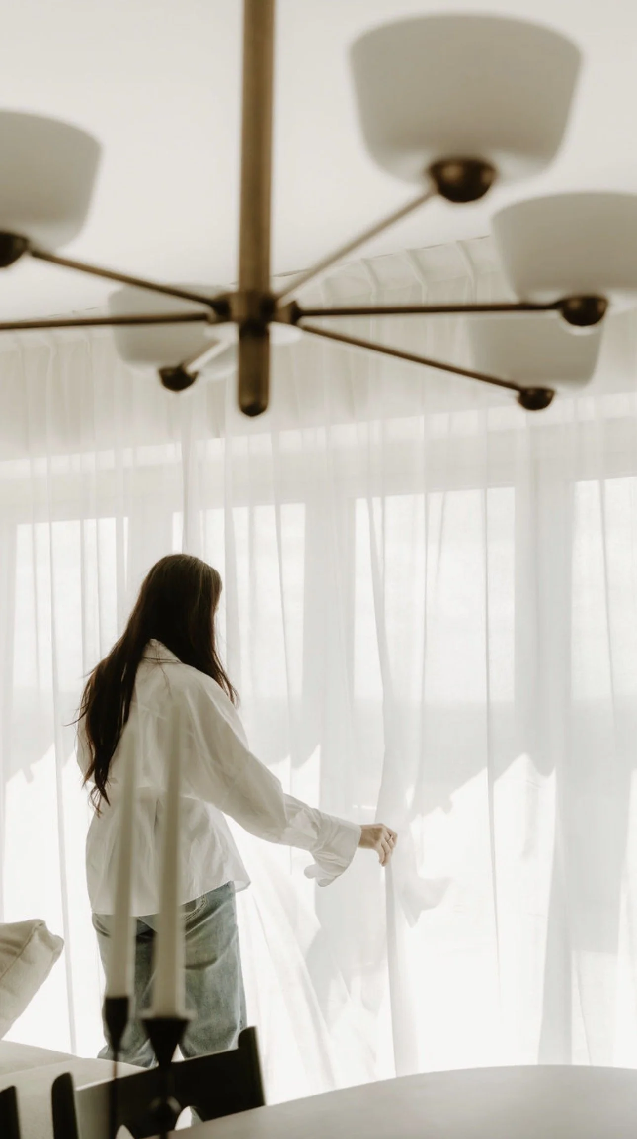 A woman in a white shirt and light-colored pants is standing near a large window with white curtains, with sunlight filtering through. The photo is taken from inside a room, possibly a kitchen or dining area, with a modern ceiling light fixture visible in the foreground.