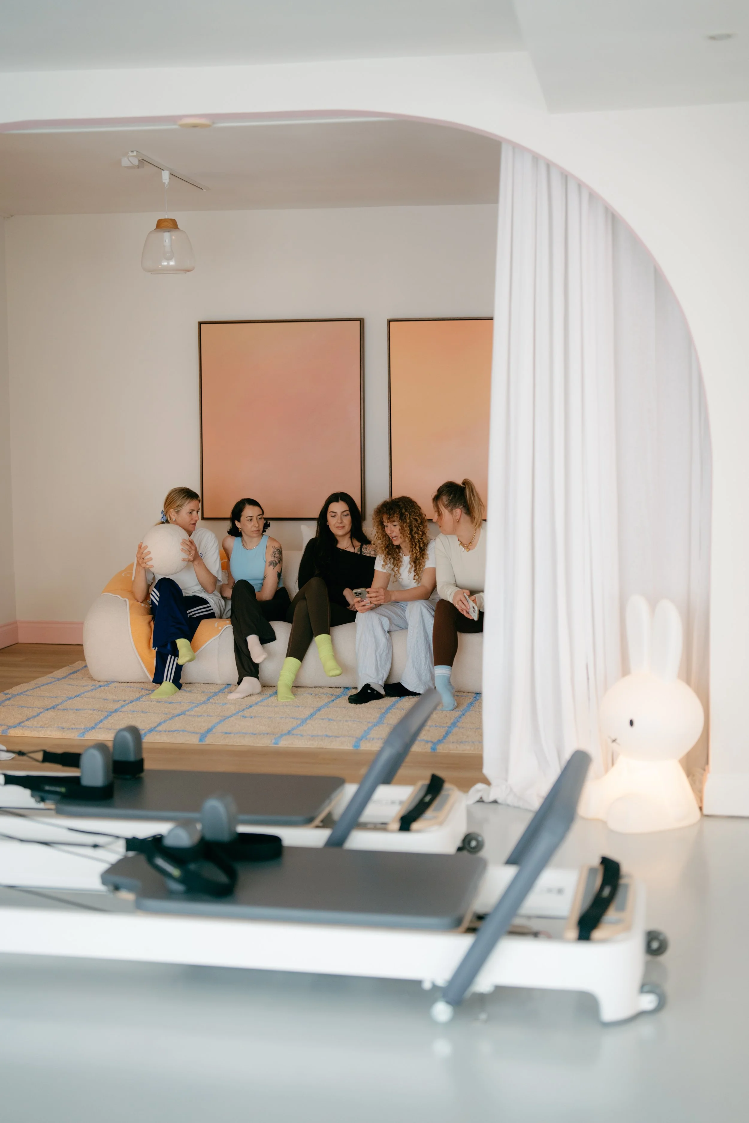 Group of five women sitting on a couch in a room with minimalist decor, two abstract paintings on the wall, and a small lamp shaped like a rabbit on a table in the foreground.