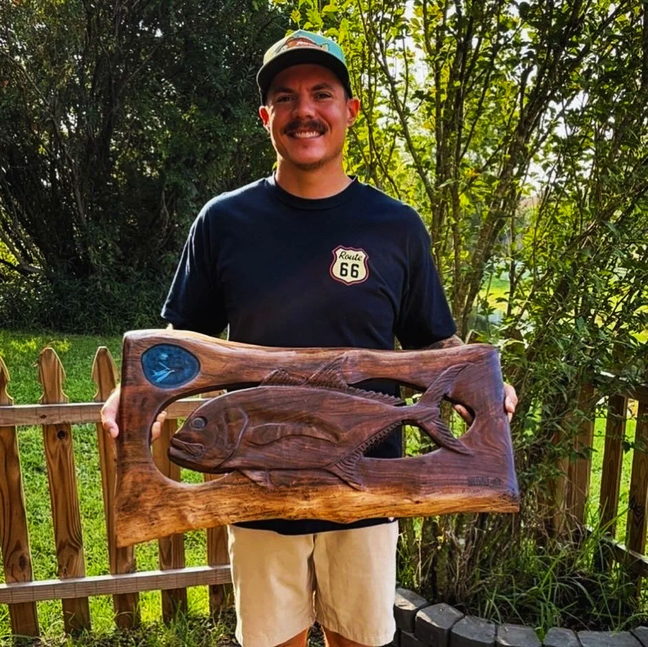 Man holding a wooden plaque with a 3D carved fish, outdoors in a backyard with trees and a wooden fence.