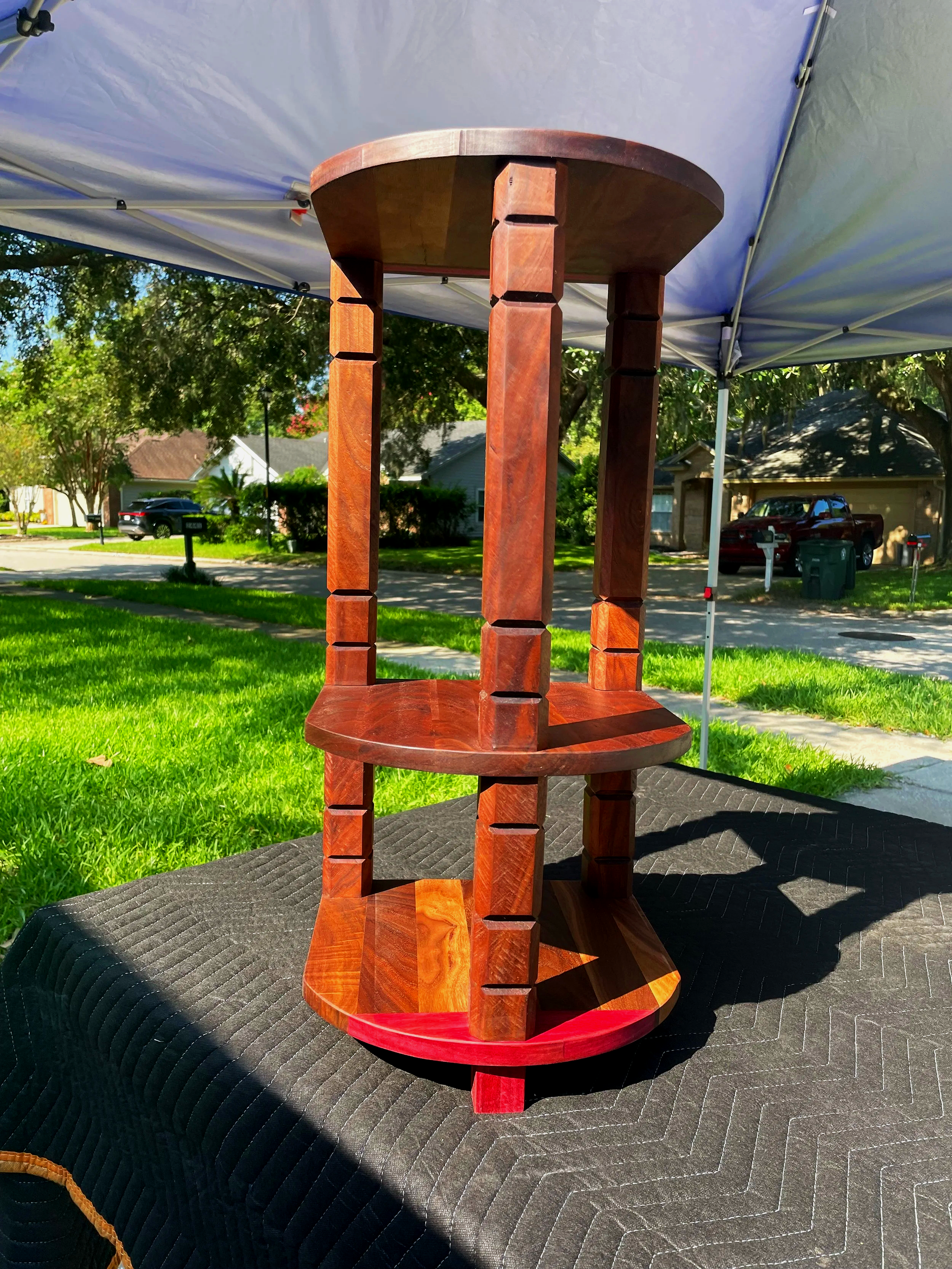 A wooden table with carved legs and a circular top, placed on a black quilted surface outdoors under a canopy. In the background, there are grass, trees, and houses.