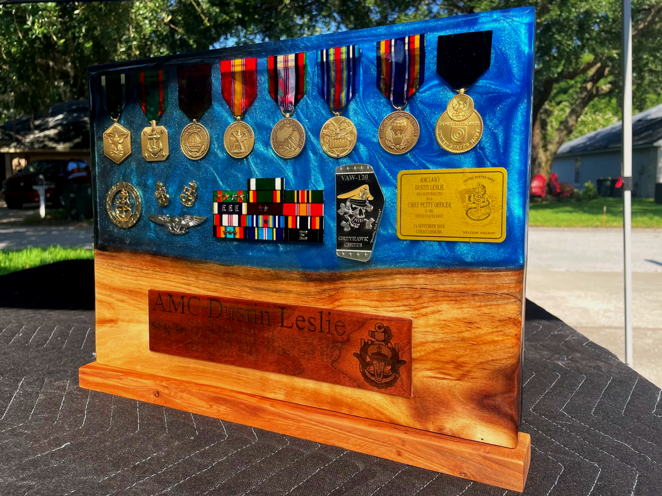 Display case with multiple military medals, ribbons, and patches on a blue background, mounted on a wooden plaque, outdoors with trees and a house in the background.
