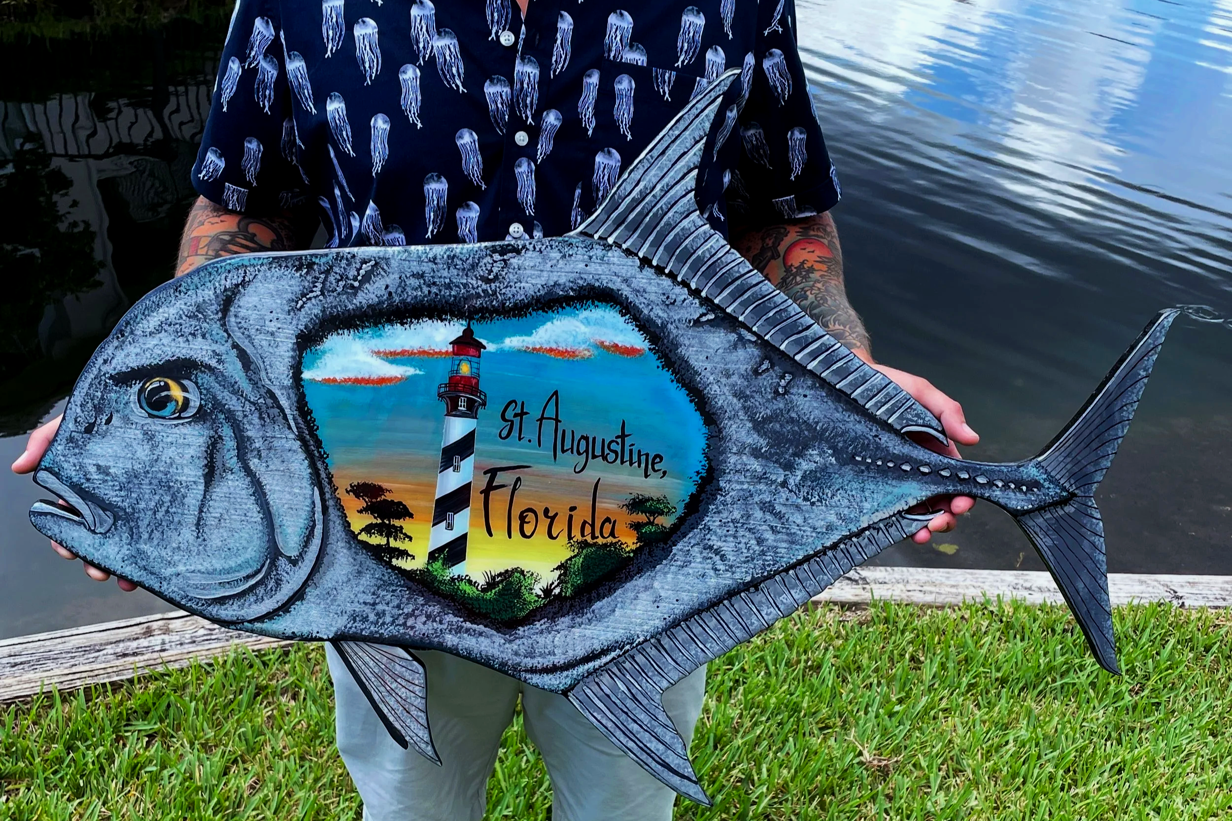 Person holding a fish-shaped sign with a lighthouse and sunset scene, reading 'St. Augustine, Florida', by a body of water, wearing a shirt with jellyfish pattern.