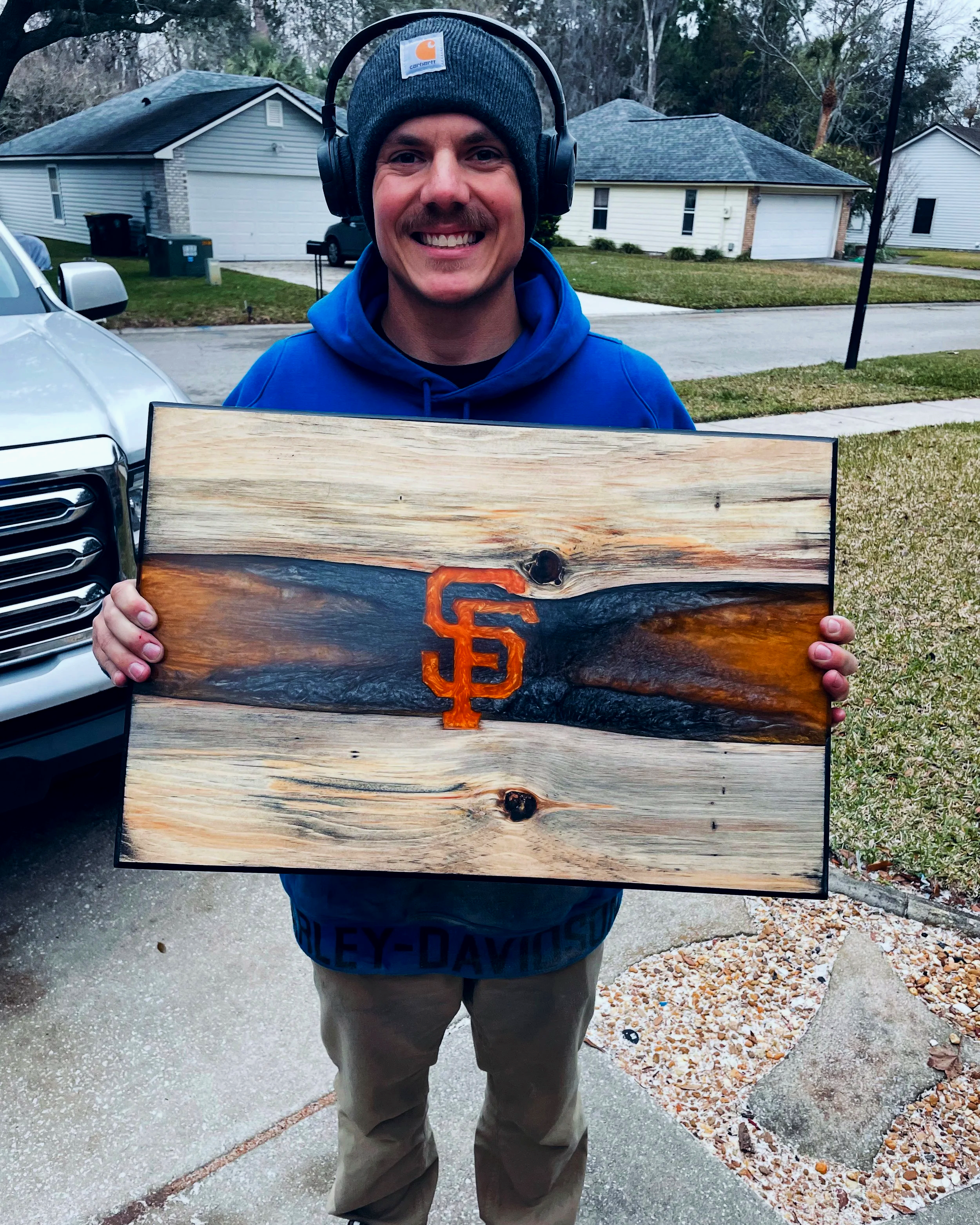 A man smiling and holding a wooden sign with the San Francisco Giants logo. He is wearing a gray Carhartt beanie and a blue hoodie. The background shows a suburban neighborhood with houses, a parked truck, and trees. There is a sidewalk and a grassy 