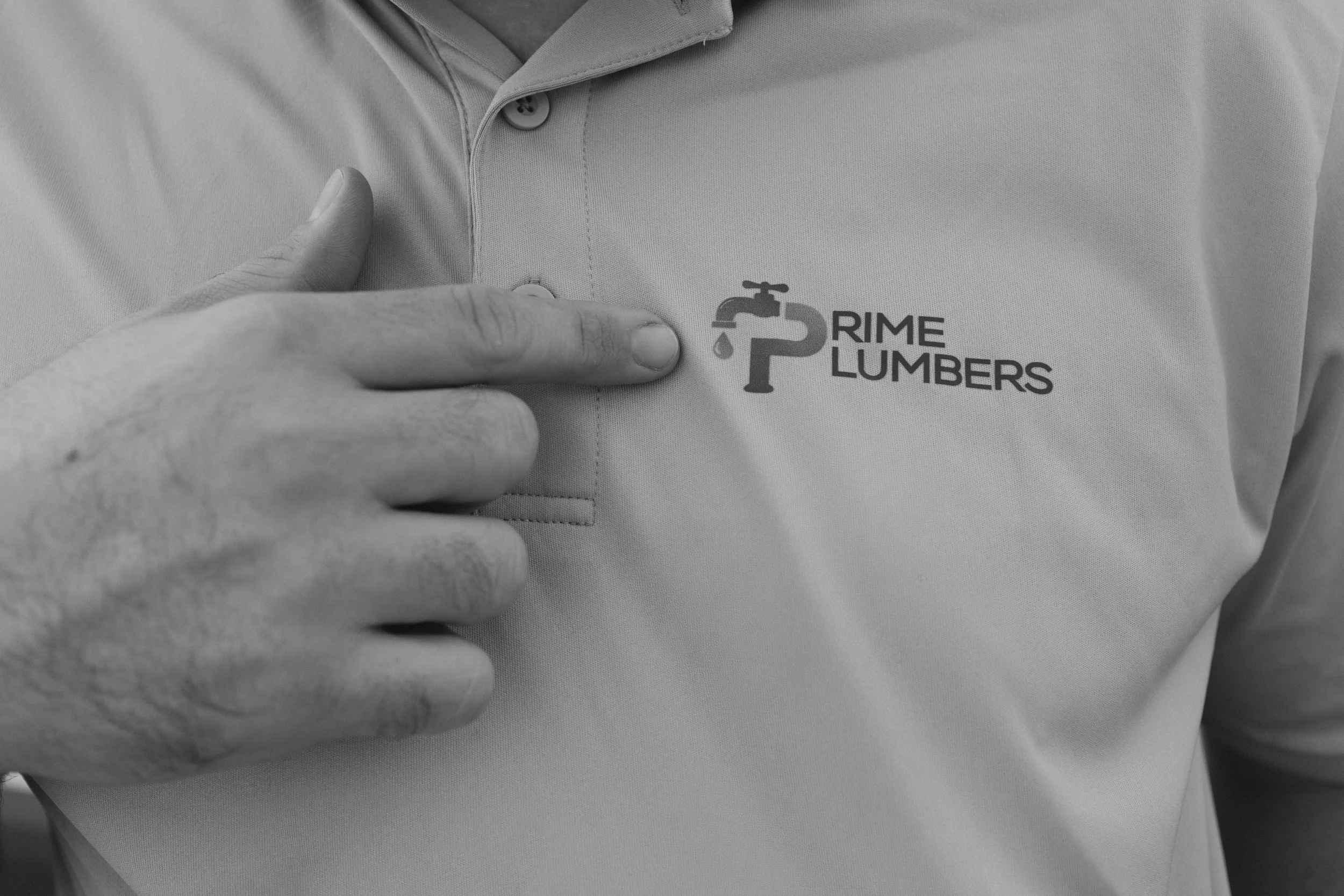 Close-up of a person pointing at a logo on their shirt. The logo includes a faucet with a droplet and the words 'Prime Plumbers'.