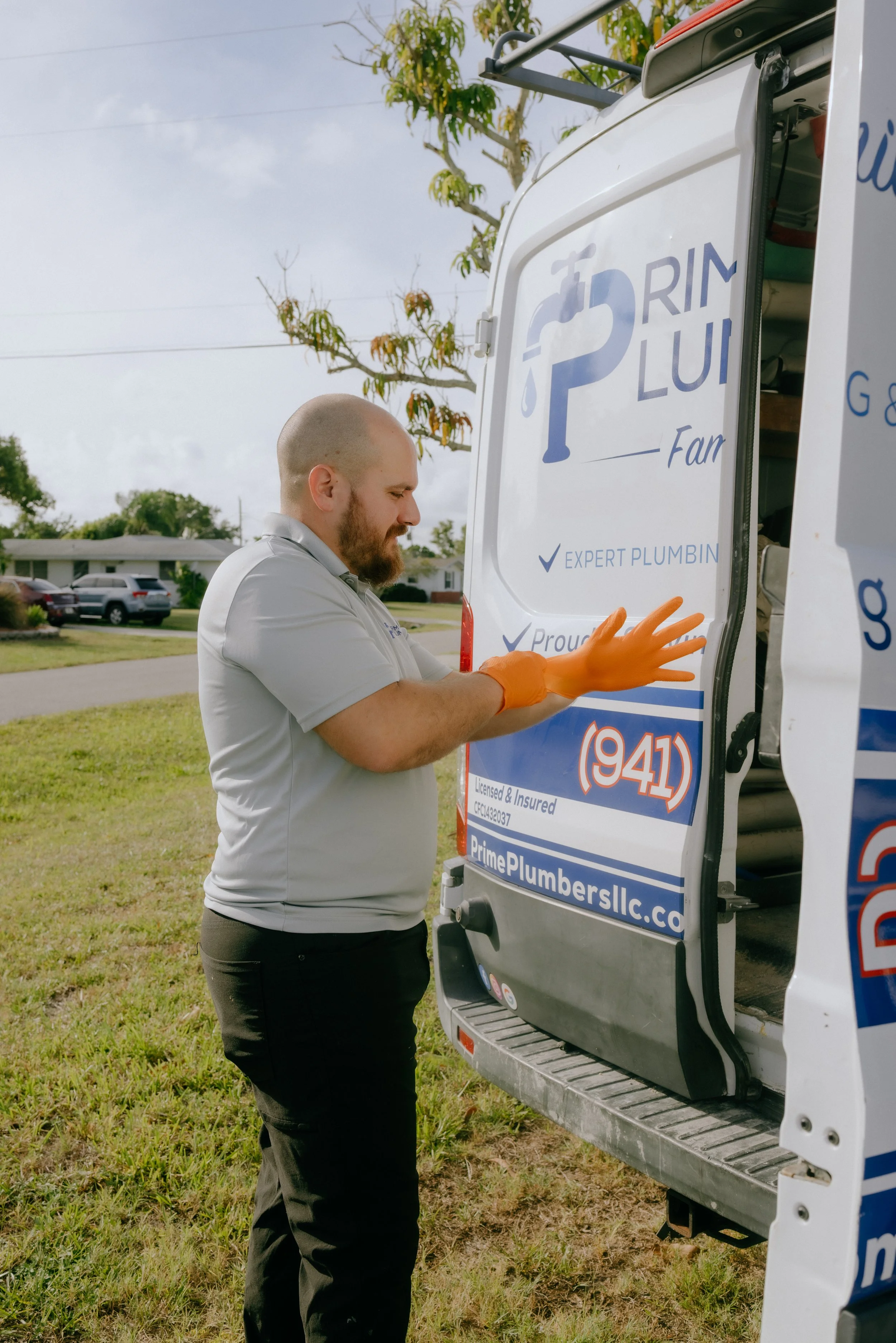 A man wearing an orange glove is standing beside a white service van with blue branding, working on the side panel at an outdoor location in a grassy area with residential houses and trees in the background.