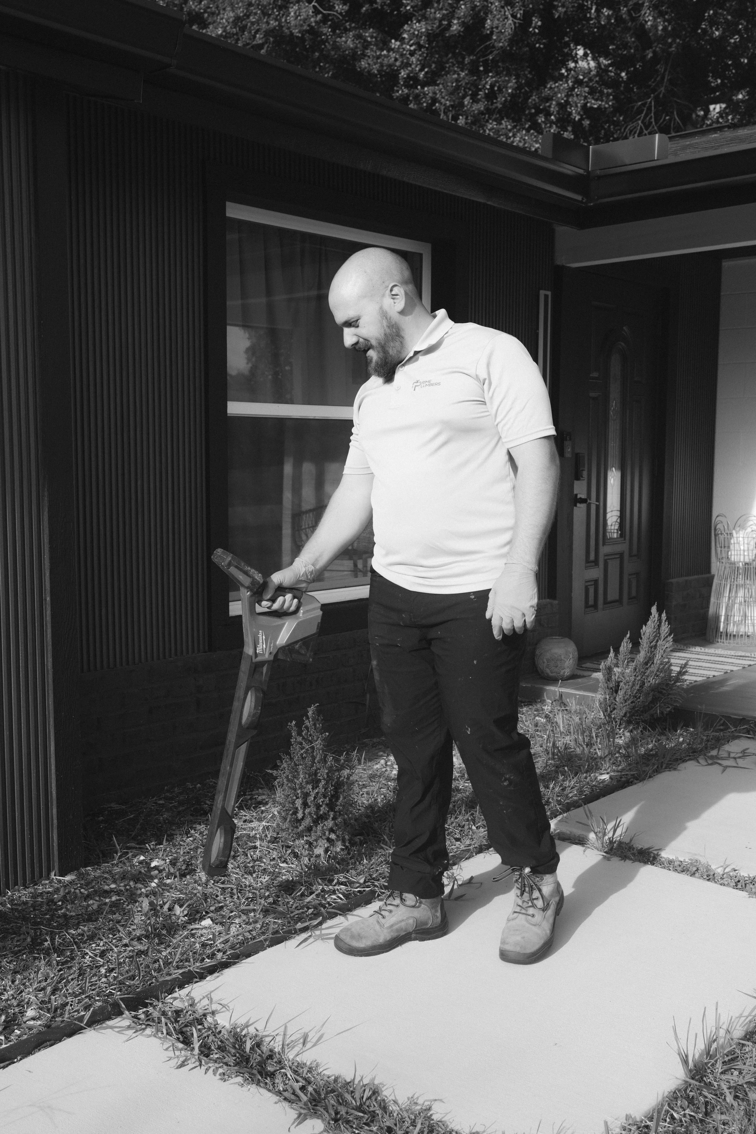 A man with a bald head, beard, and light-colored polo shirt stands outside on a concrete walkway, looking down at an object in his hands. He is wearing dark pants and work boots. Behind him is a house with dark siding, a large window, and a door, along with some plants and garden decorations.