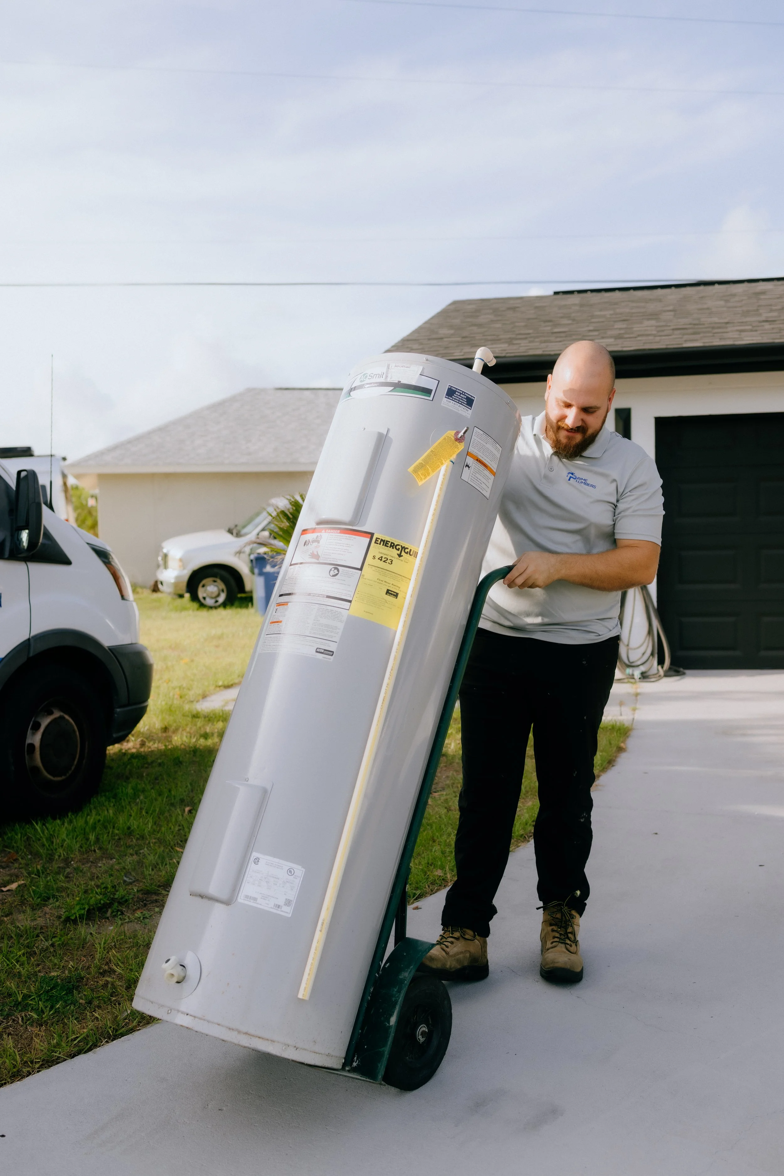 A man in work boots and a gray shirt loading a water heater onto a dolly in a residential driveway.