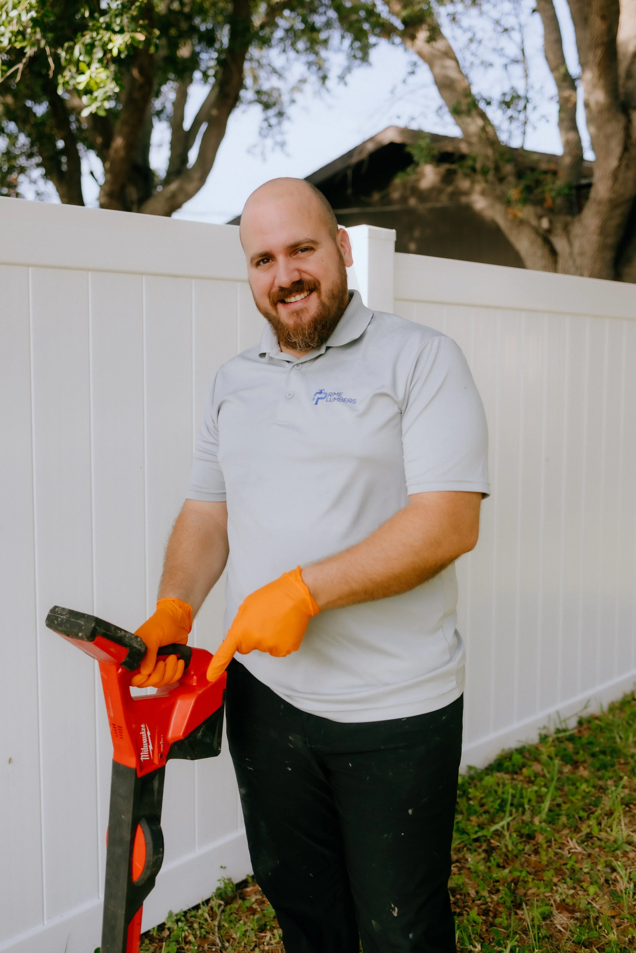 Man with a beard wearing a gray polo shirt with a logo, orange gloves, and black pants, working with a handheld power tool outdoors, standing next to a white fence and surrounded by trees and grass.