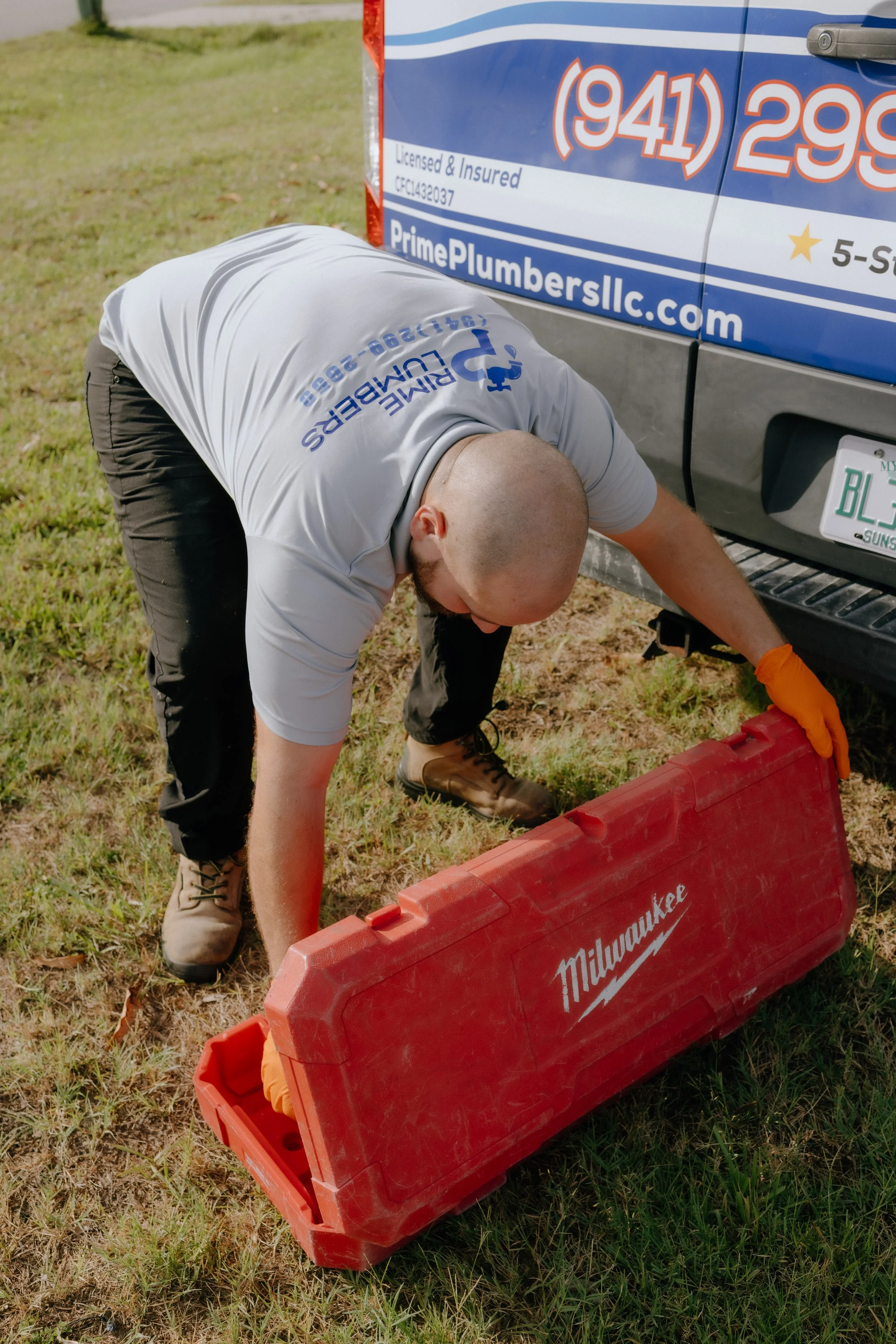A man wearing a white shirt with blue text and black pants is kneeling on grass, working on something near a red Milwaukee toolbox. The toolbox is open, revealing some tools, and is placed on the ground beside a blue service van with company information and phone number.