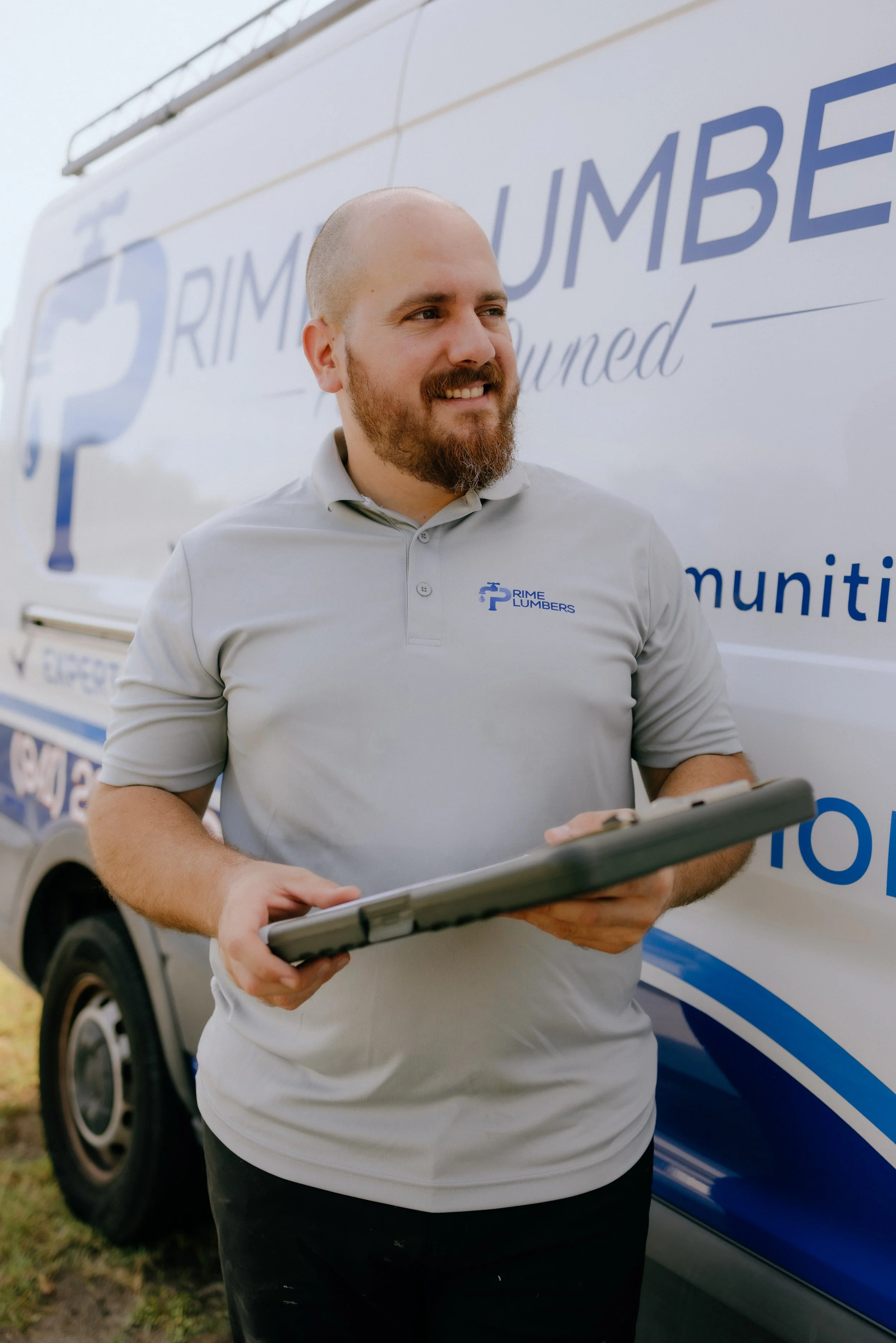 A man with a beard holding a tablet in front of a vehicle with a sign reading "RIME LUMBERS" and a star logo, wearing a light-colored polo shirt.