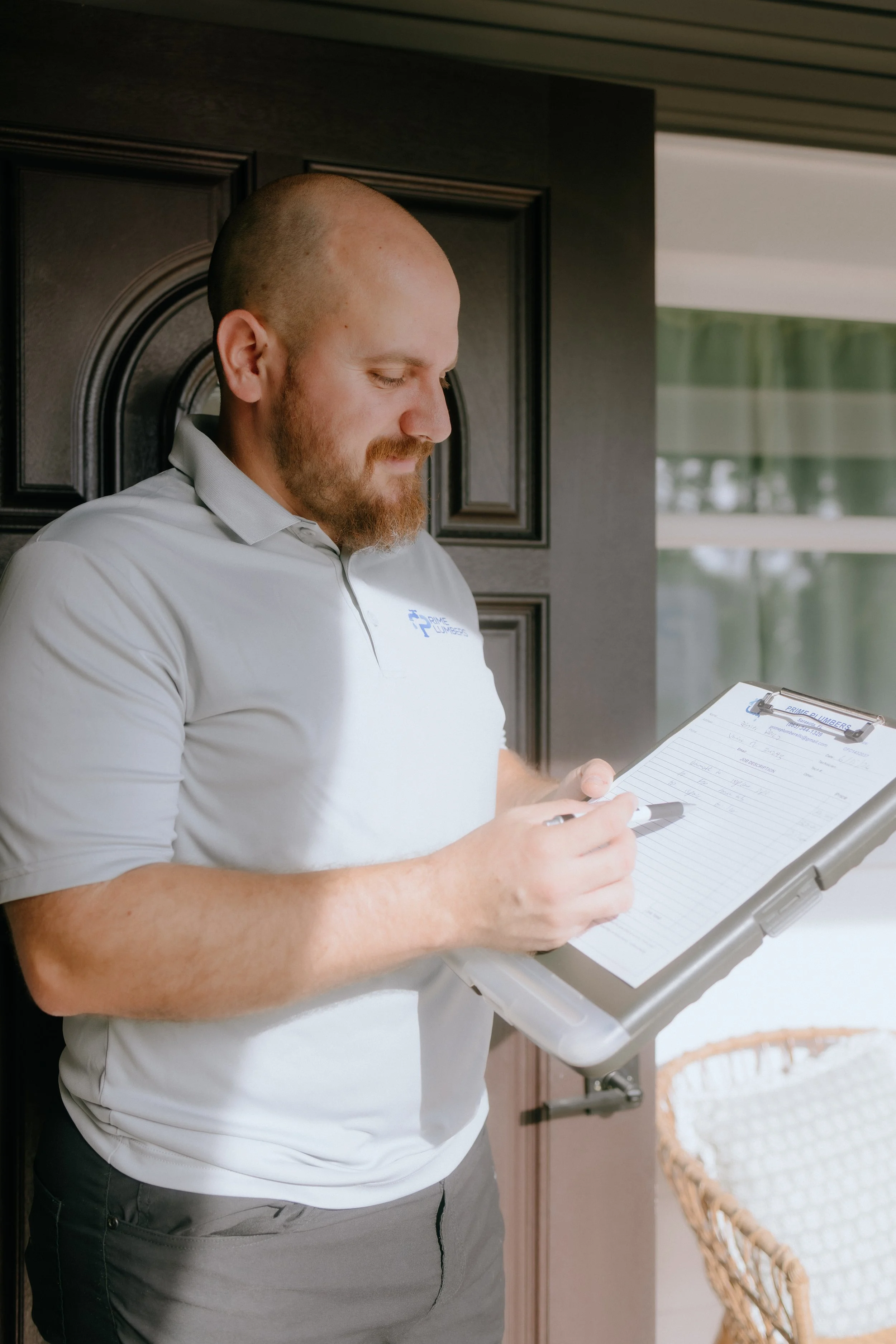 A man with a beard and a shaved head, wearing a light grey polo shirt and grey pants, is standing indoors holding a clipboard and writing with a pen, with a window and wicker chair in the background.