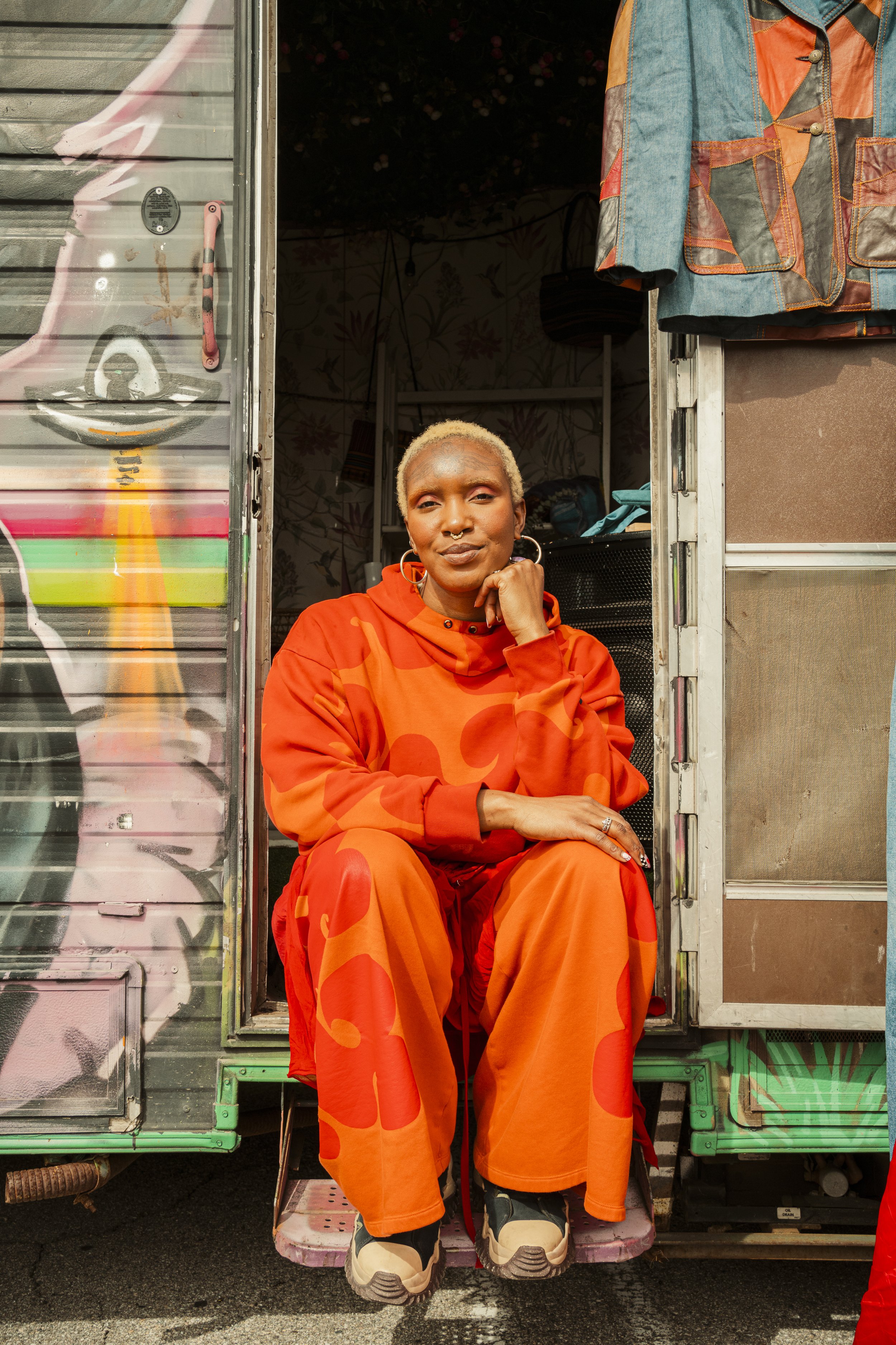 A person with short platinum blonde hair and large hoop earrings sitting on the edge of a mobile trailer, wearing an orange camo-patterned outfit and chunky black and beige sneakers.
