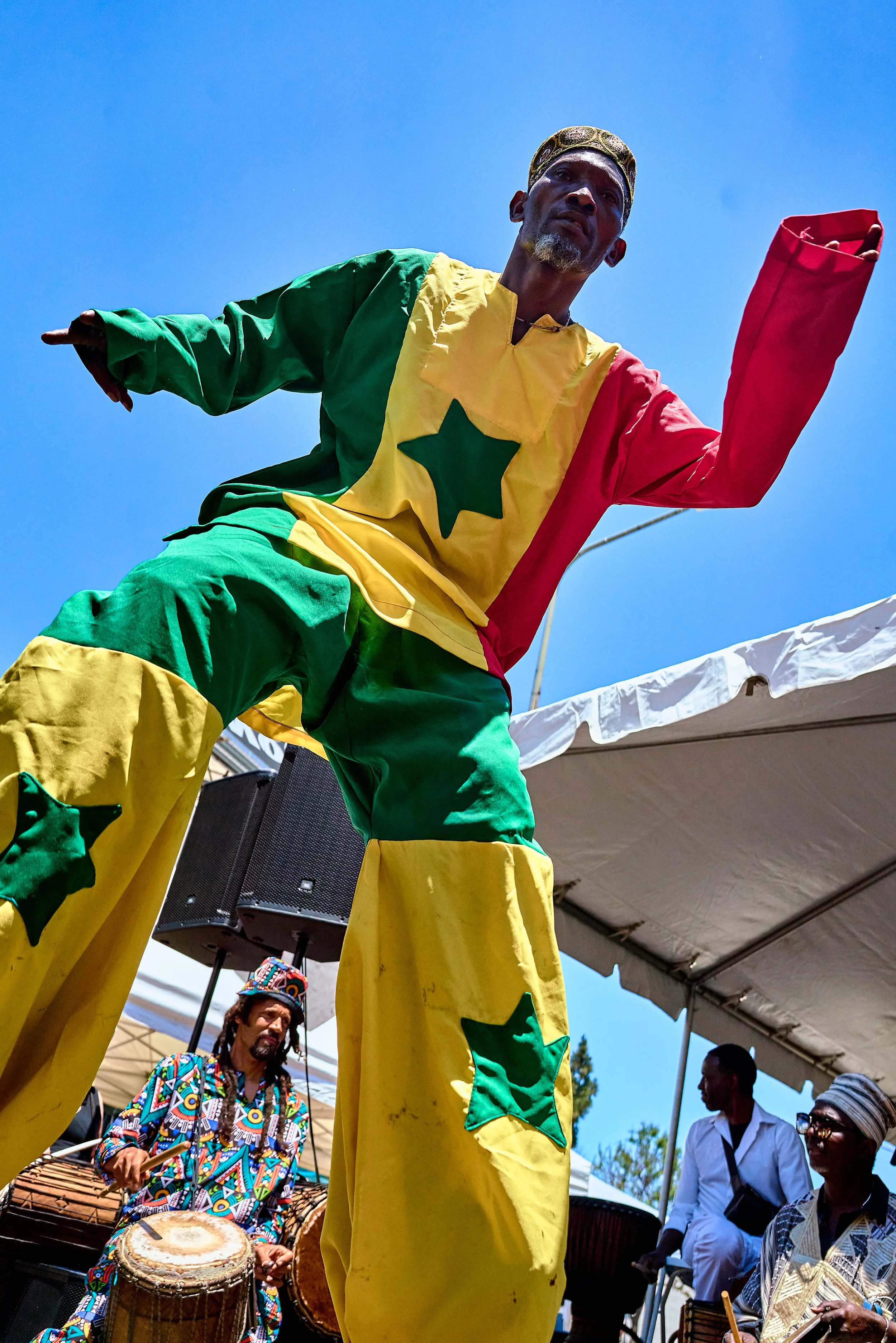 A stilt performer dressed in colorful African attire with a yellow, green, and red outfit and star designs, is dancing with other musicians and performers, including a man playing drums, under a bright blue sky and a white tent.