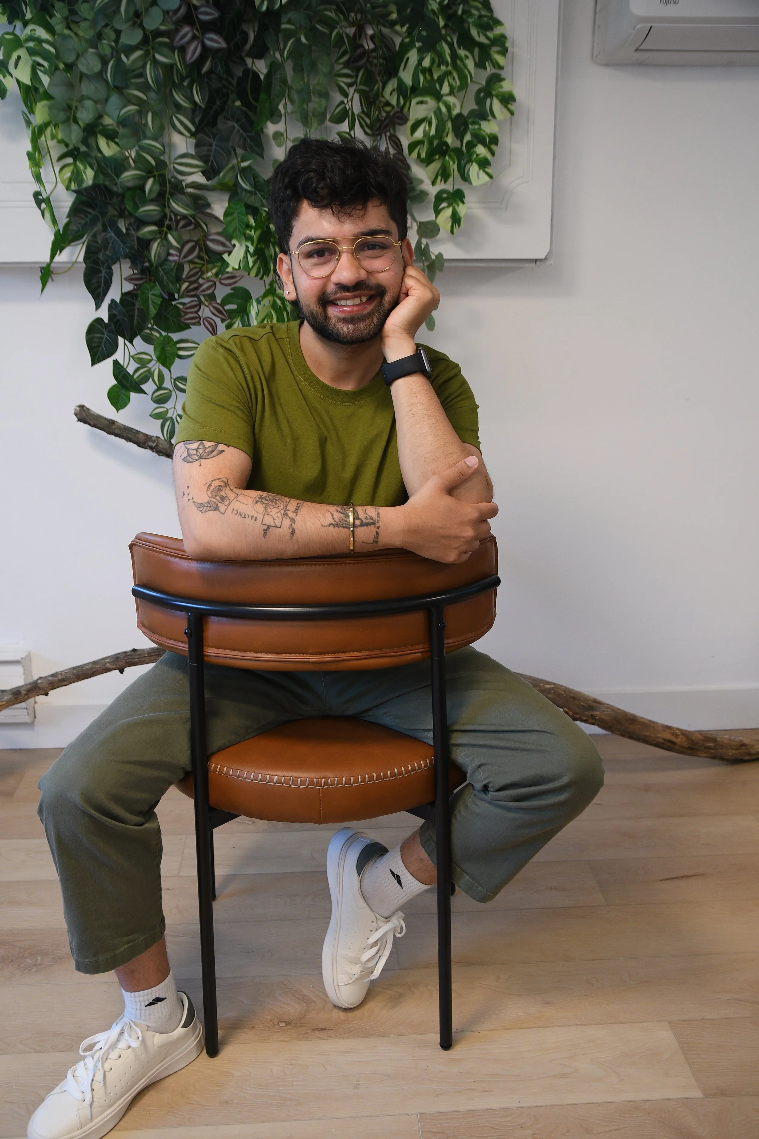 A young man with dark curly hair, glasses, and a beard, wearing a green t-shirt and gray pants, sitting on a chair with a leather back, smiling with his head resting on his hand. He has tattoos on his arms, and is wearing white sneakers and a black watch. Behind him is a plant with variegated leaves, and a white wall with an air conditioning unit.