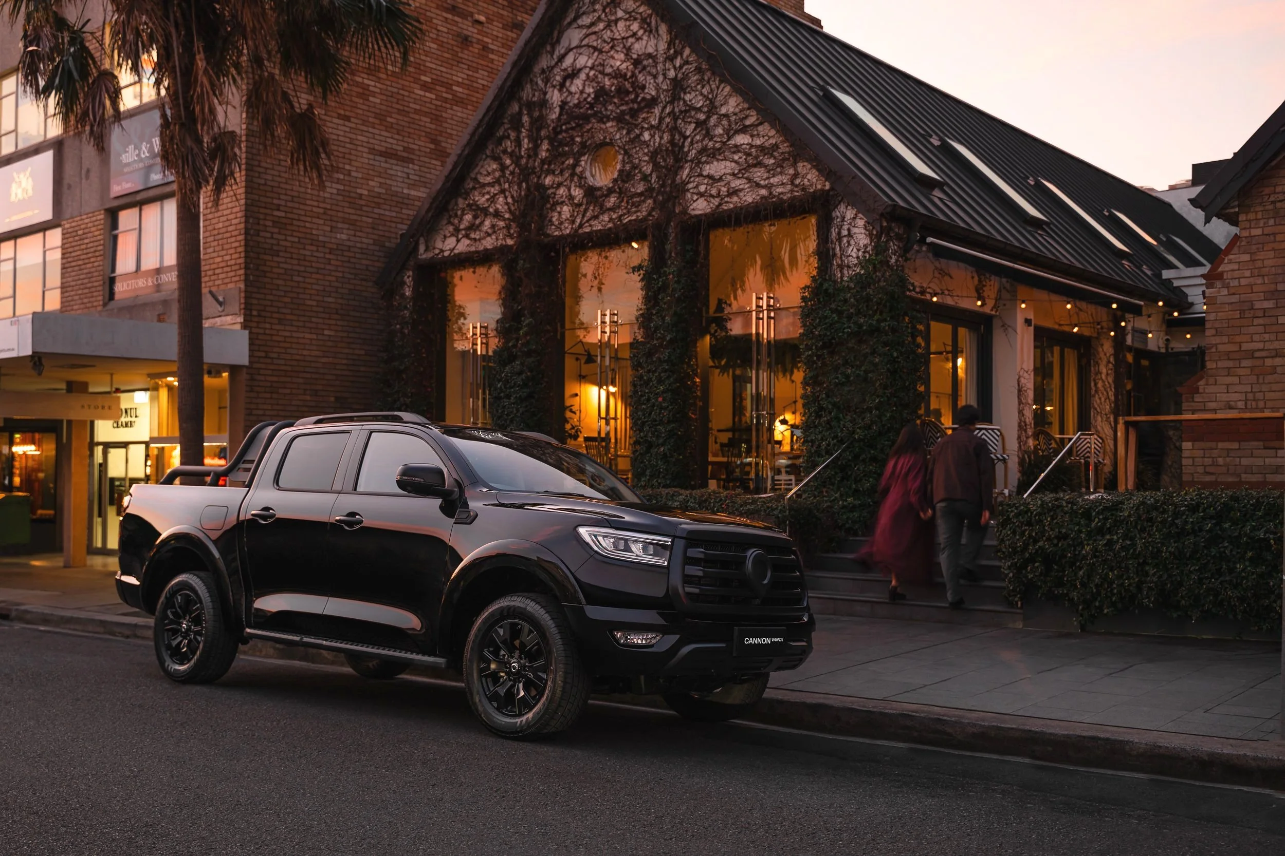 A black pickup truck parked on the street in front of a restaurant with large windows and string lights. The restaurant has a brick and glass exterior with vines growing on part of the building. There are two people walking past the restaurant, and t