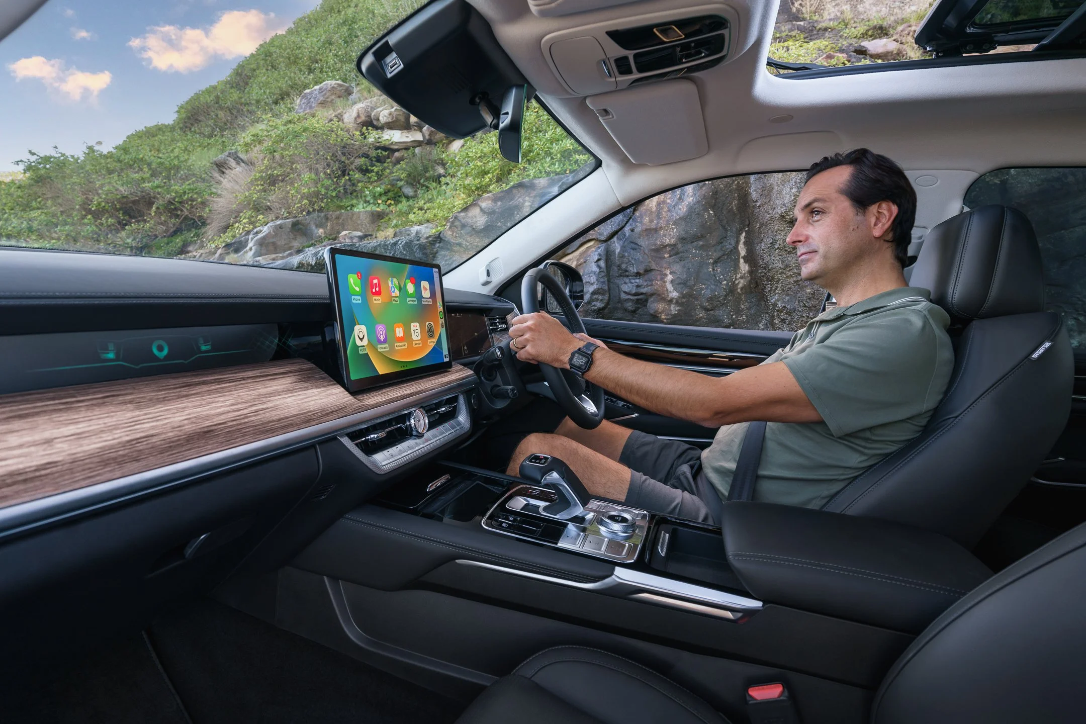 A man driving a vehicle with a digital display screen showing app icons, with a rocky, green outdoor landscape visible through the windshield.