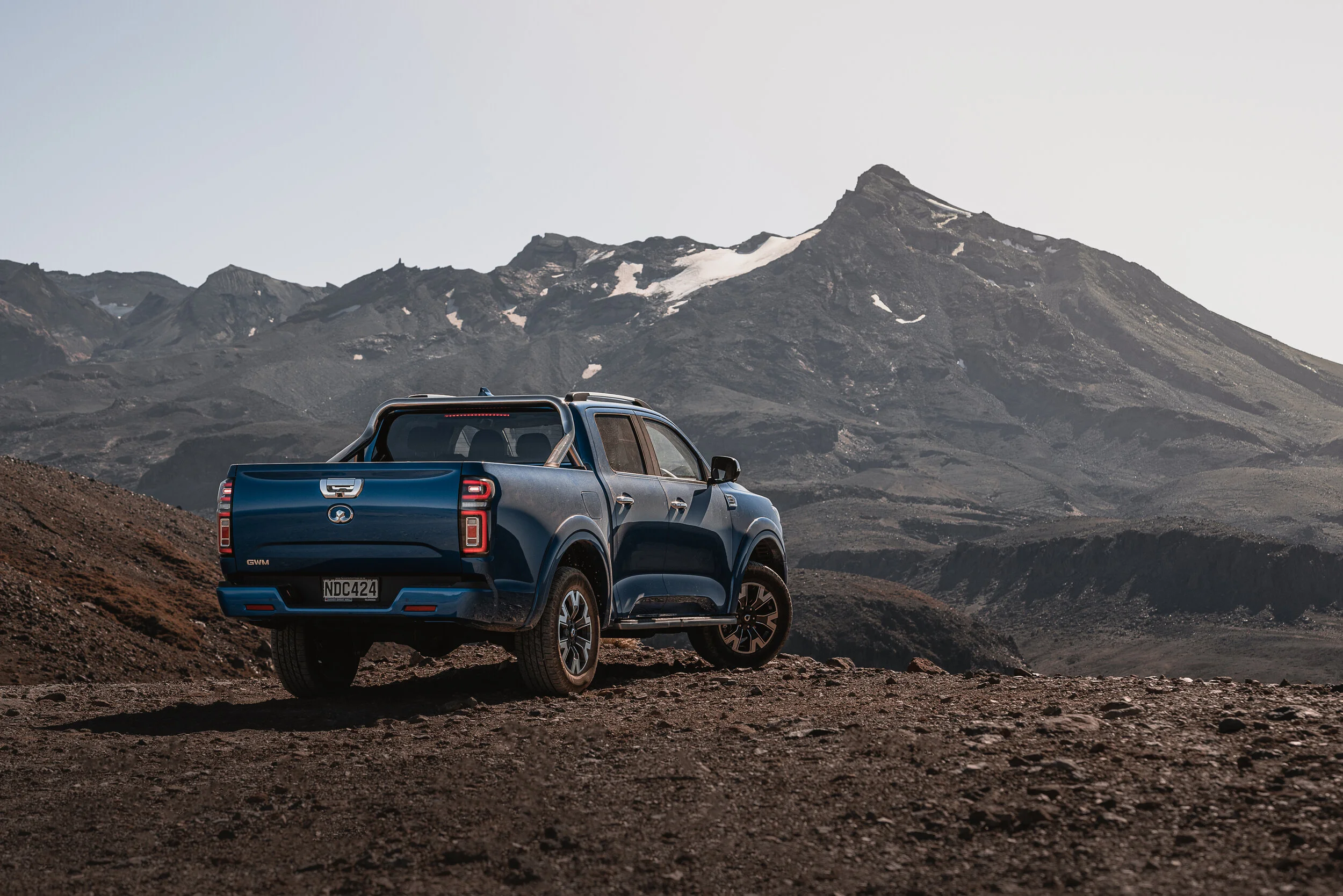 A blue pickup truck parked on rocky terrain with a mountain and snow patches in the background.
