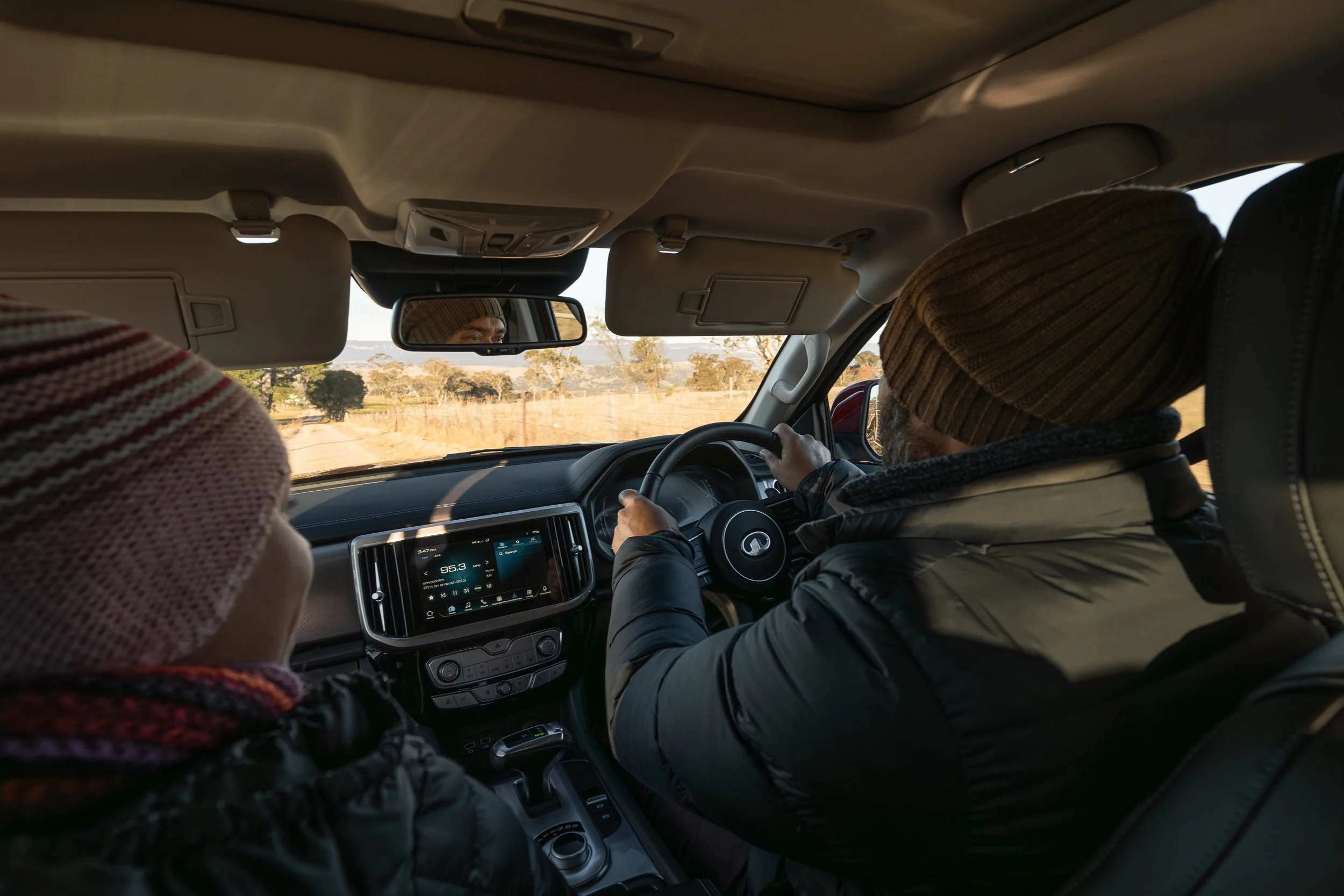 Interior of a vehicle with two passengers, one driving and one in the passenger seat, both wearing knit hats, on a rural road with open fields and trees visible through the windshield.