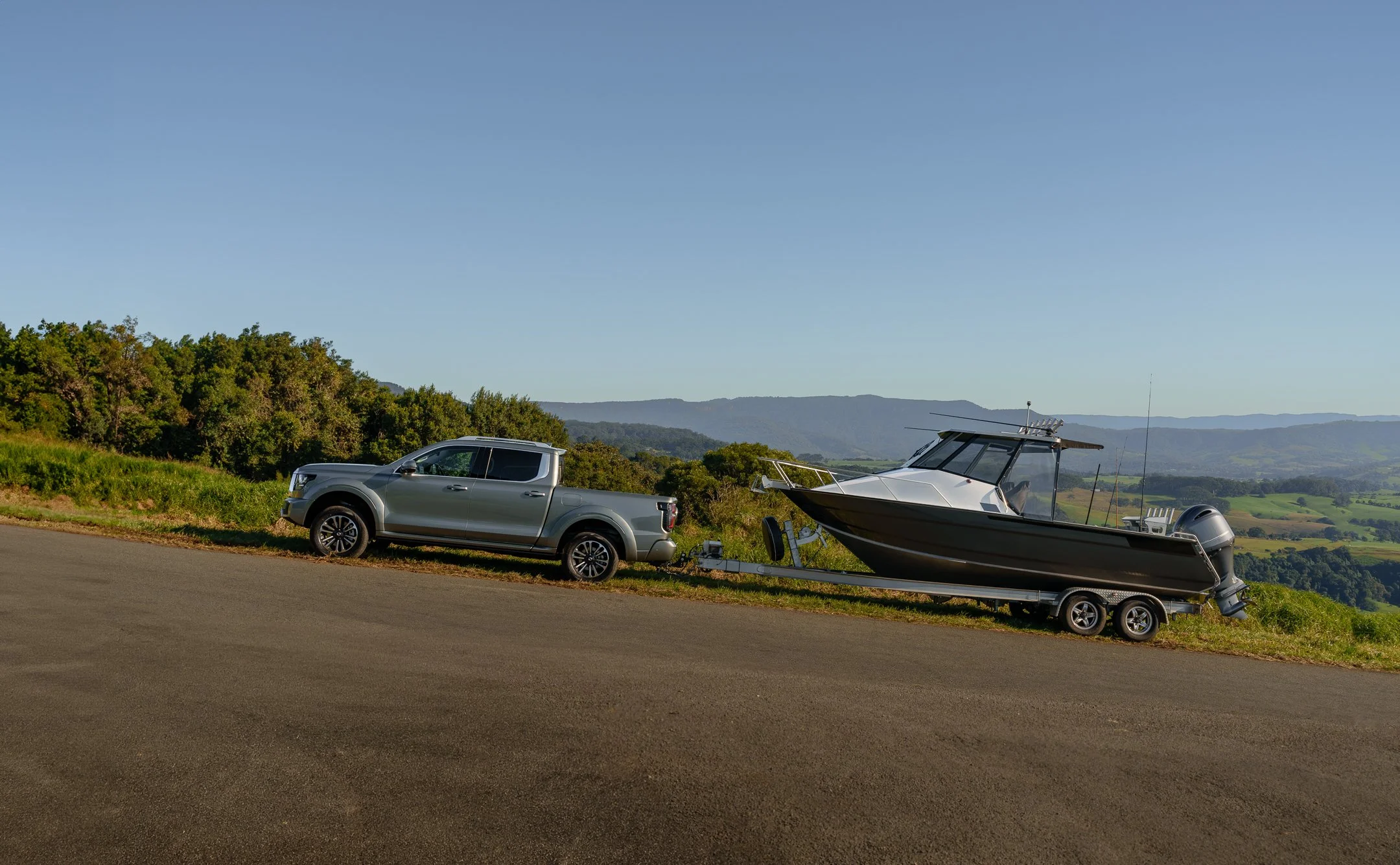 A pickup truck towing a boat on a trailer parked on the side of a road with green trees and hills in the background under a clear blue sky.