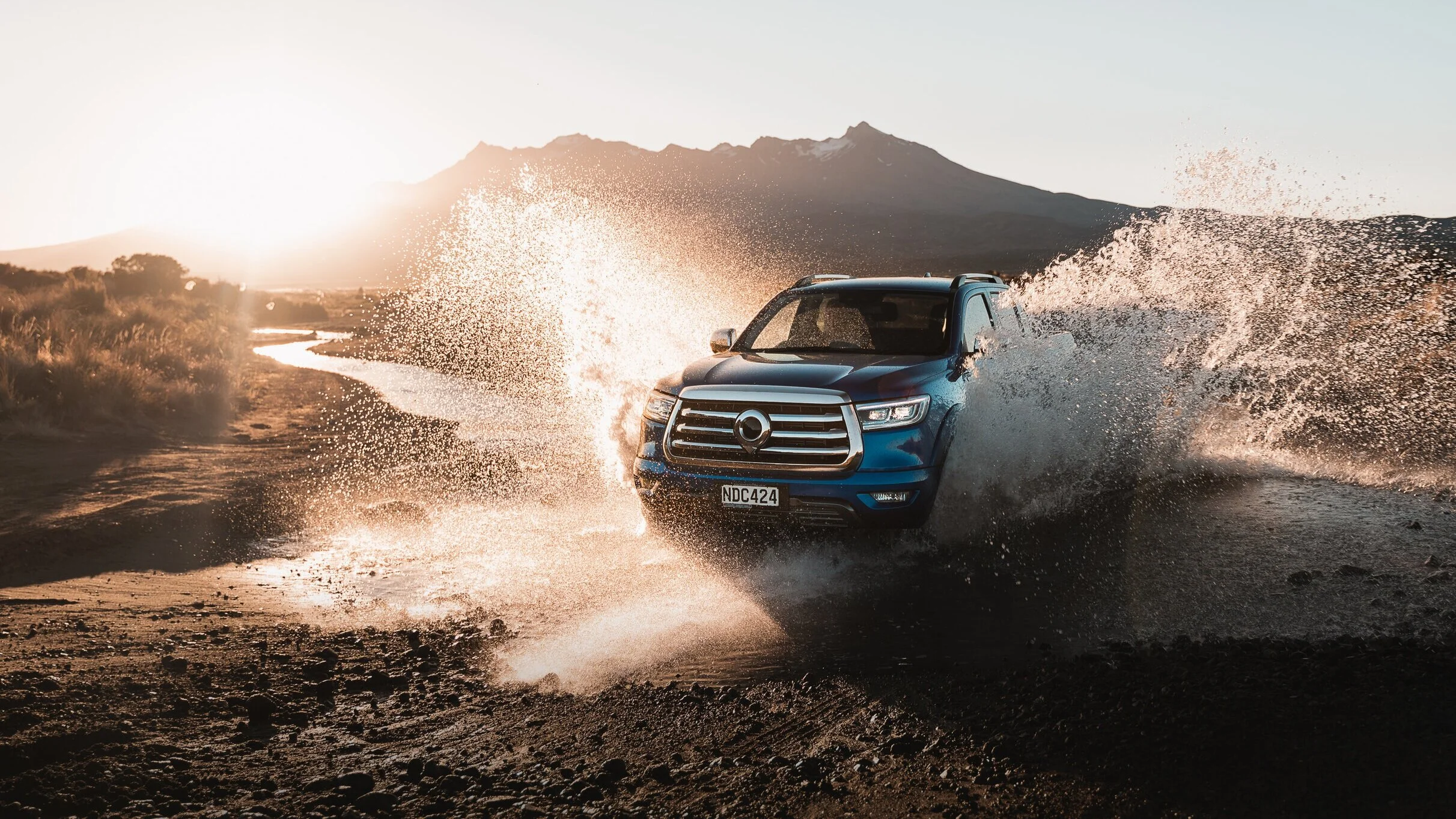 A blue SUV driving through a flooded dirt road with mountains in the background during sunset.