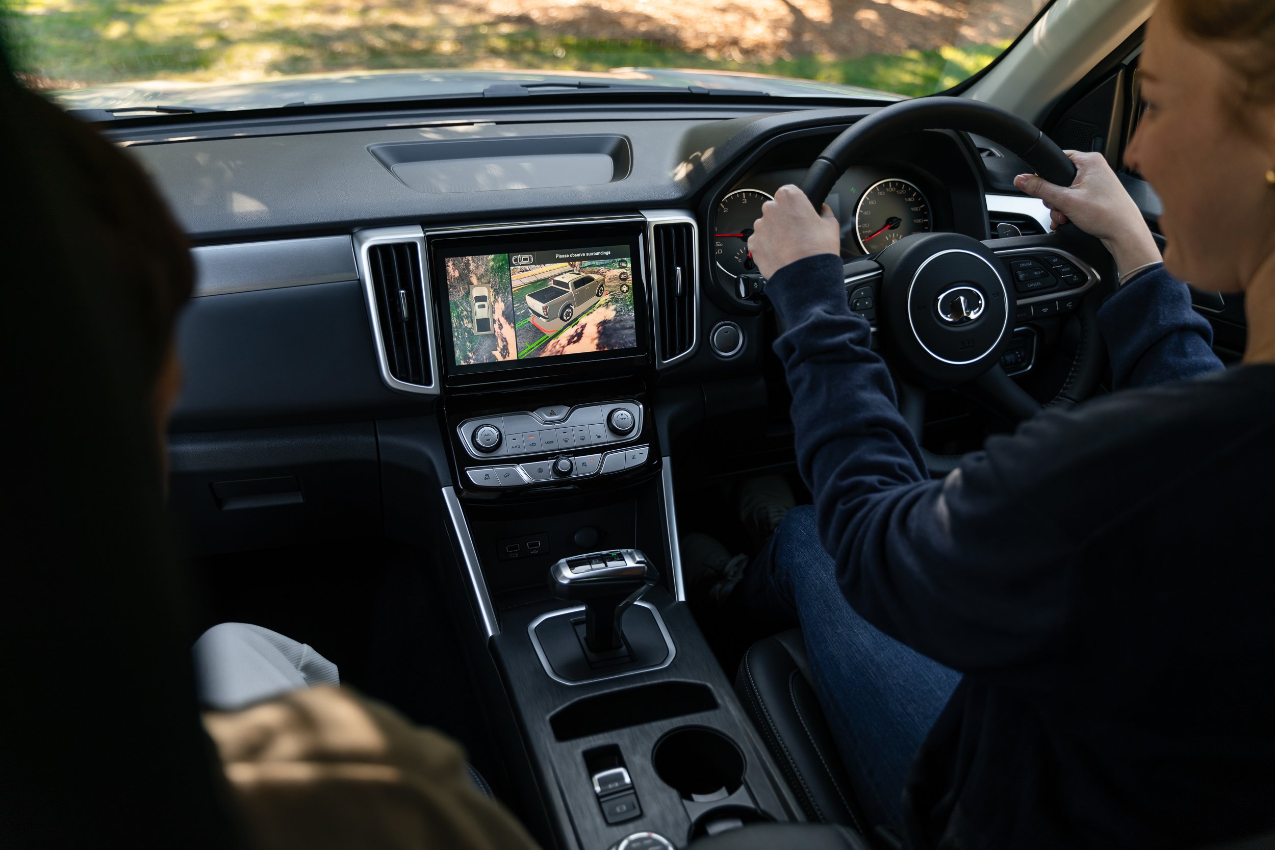 Inside view of a vehicle with a person driving and a passenger, showing the dashboard and a display screen with a vehicle surroundings view.