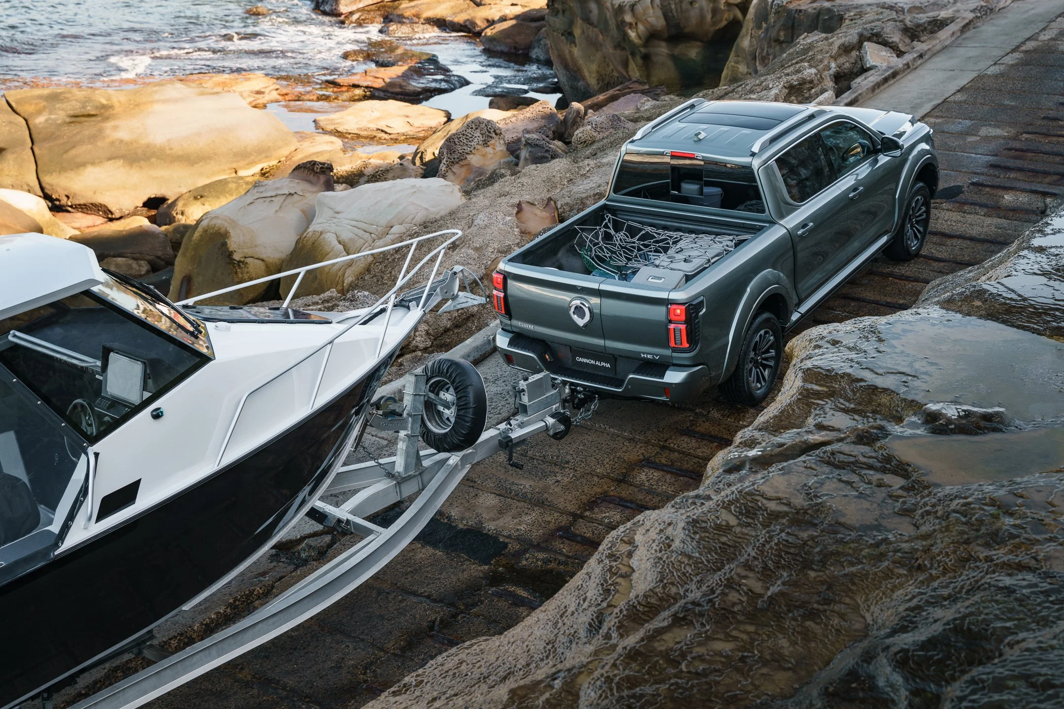 A pickup truck with a boat on a trailer parked near a rocky shoreline by the water.