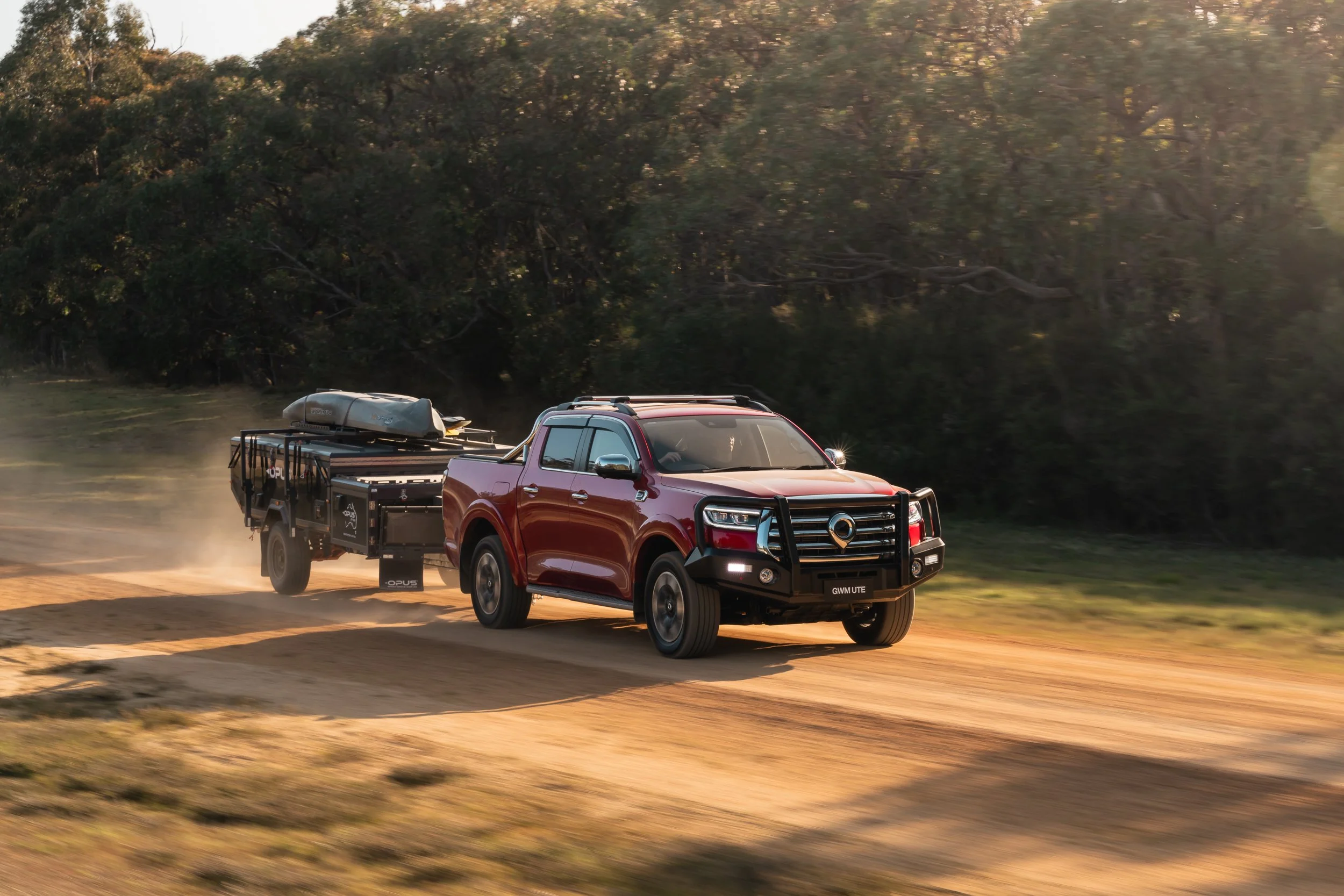 A red pickup truck with black off-road accessories driving on a dirt road, kicking up dust, with a background of trees and sunlight filtering through the foliage.