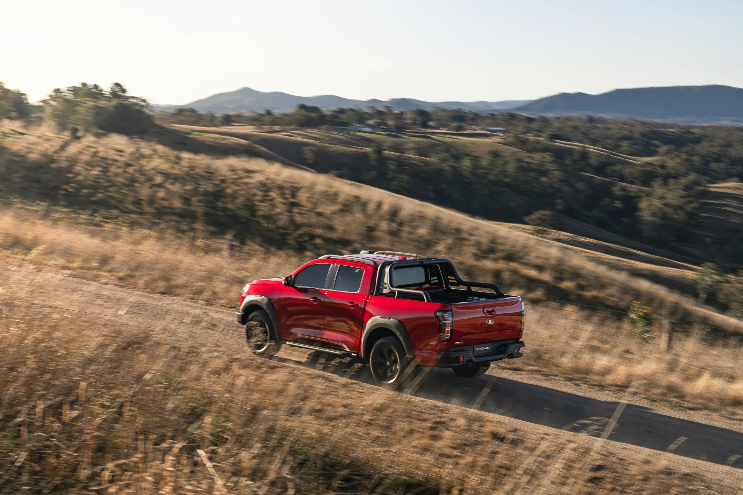 Red pickup truck driving on a dirt road through rural hills with mountains in the background.