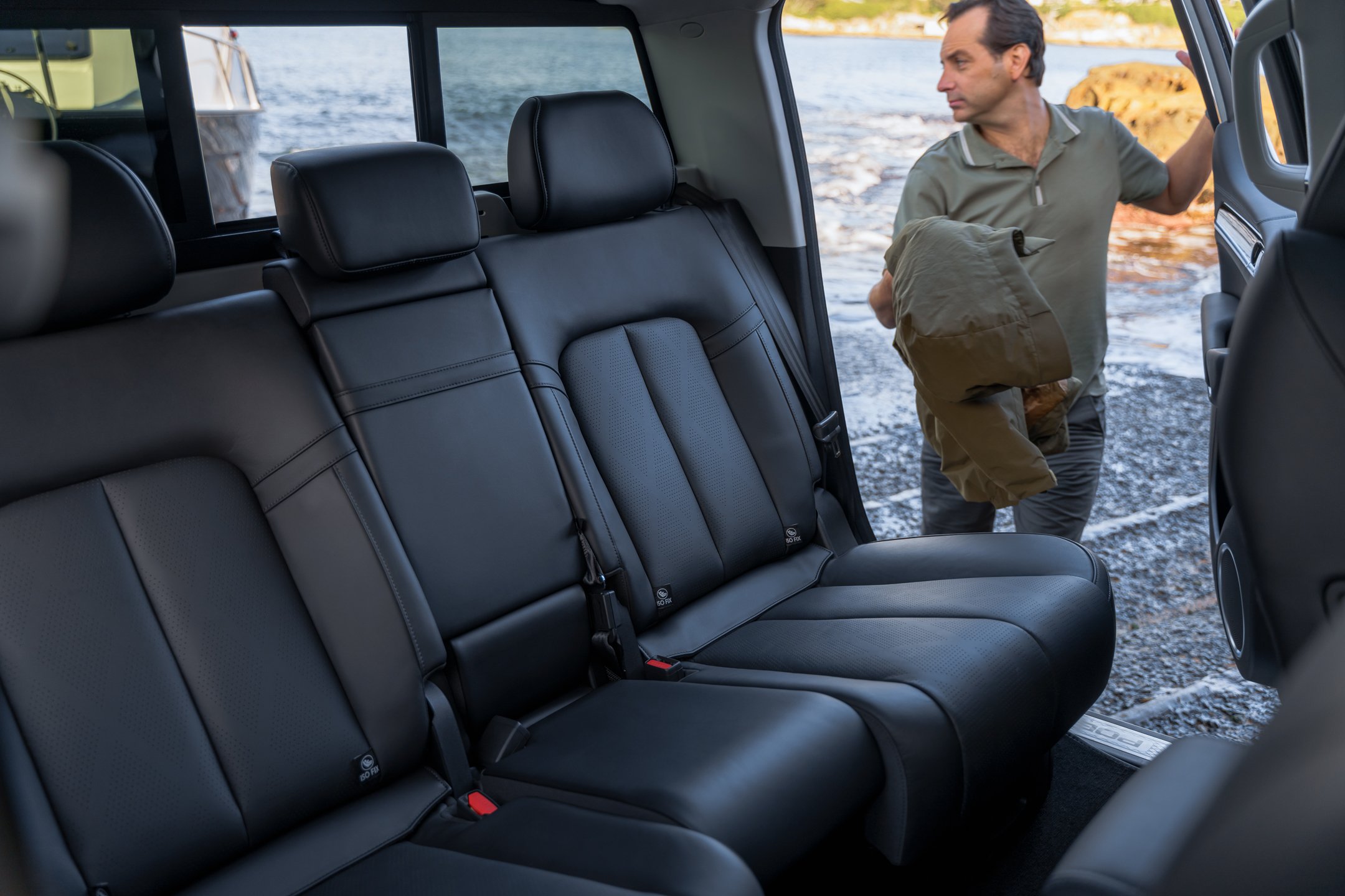 Interior view of a black leather vehicle seat with an outdoor scene visible through the window, where a man is standing near the water holding a brown bag and looking to the right.