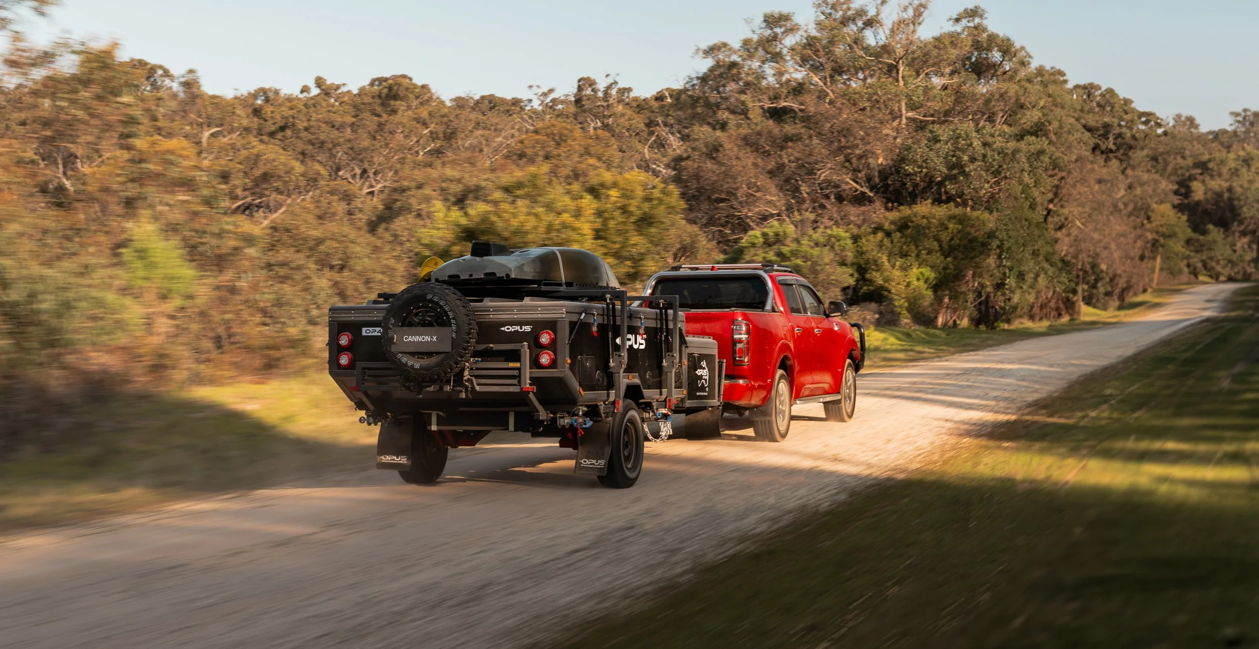A red pickup truck with a trailer attached, driving on a dirt road through a wooded area, with trees and bushes on either side.