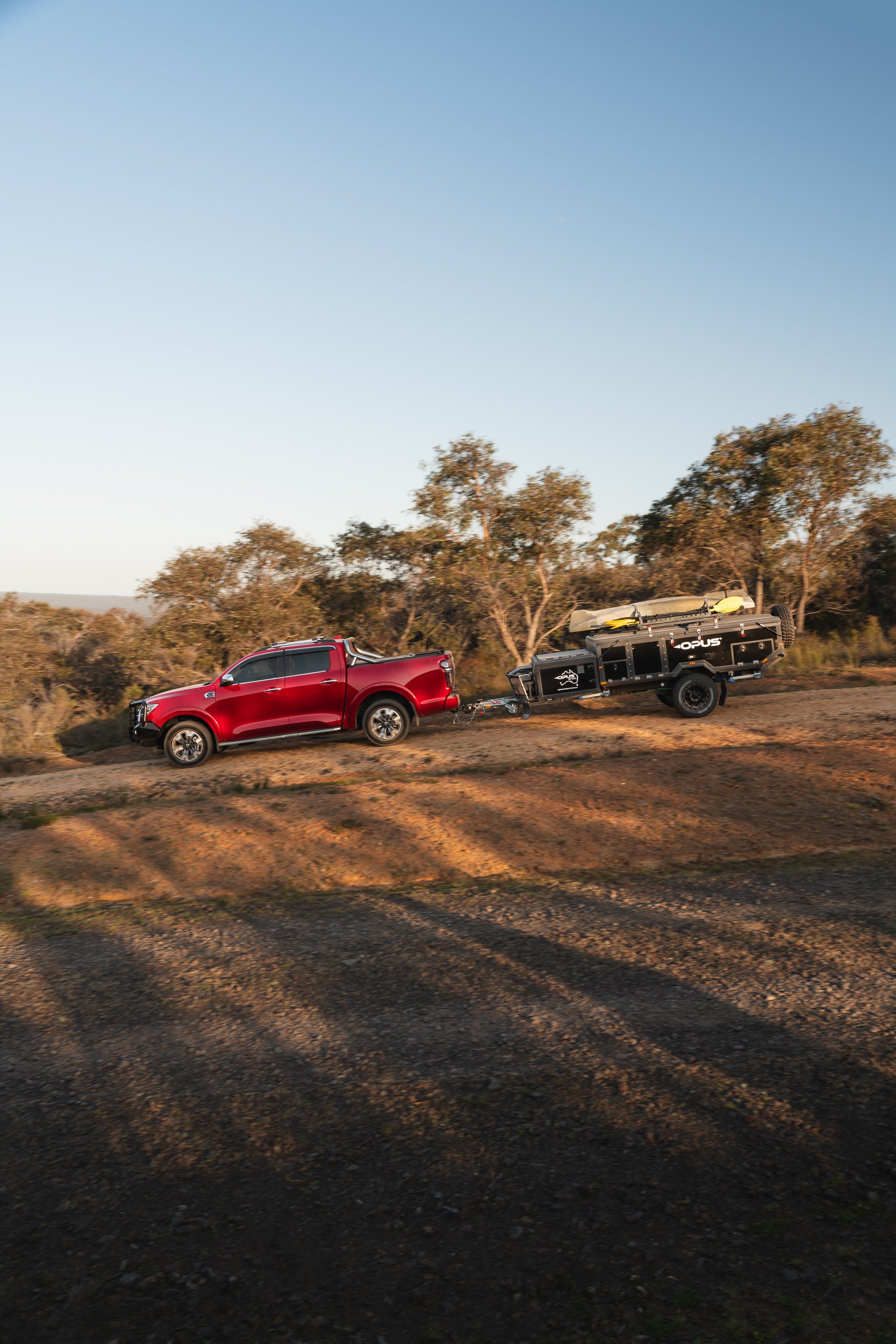 A red pickup truck hauling a black off-road camper trailer with a kayak on top, traveling on a dirt road through a landscape with trees and a clear sky.