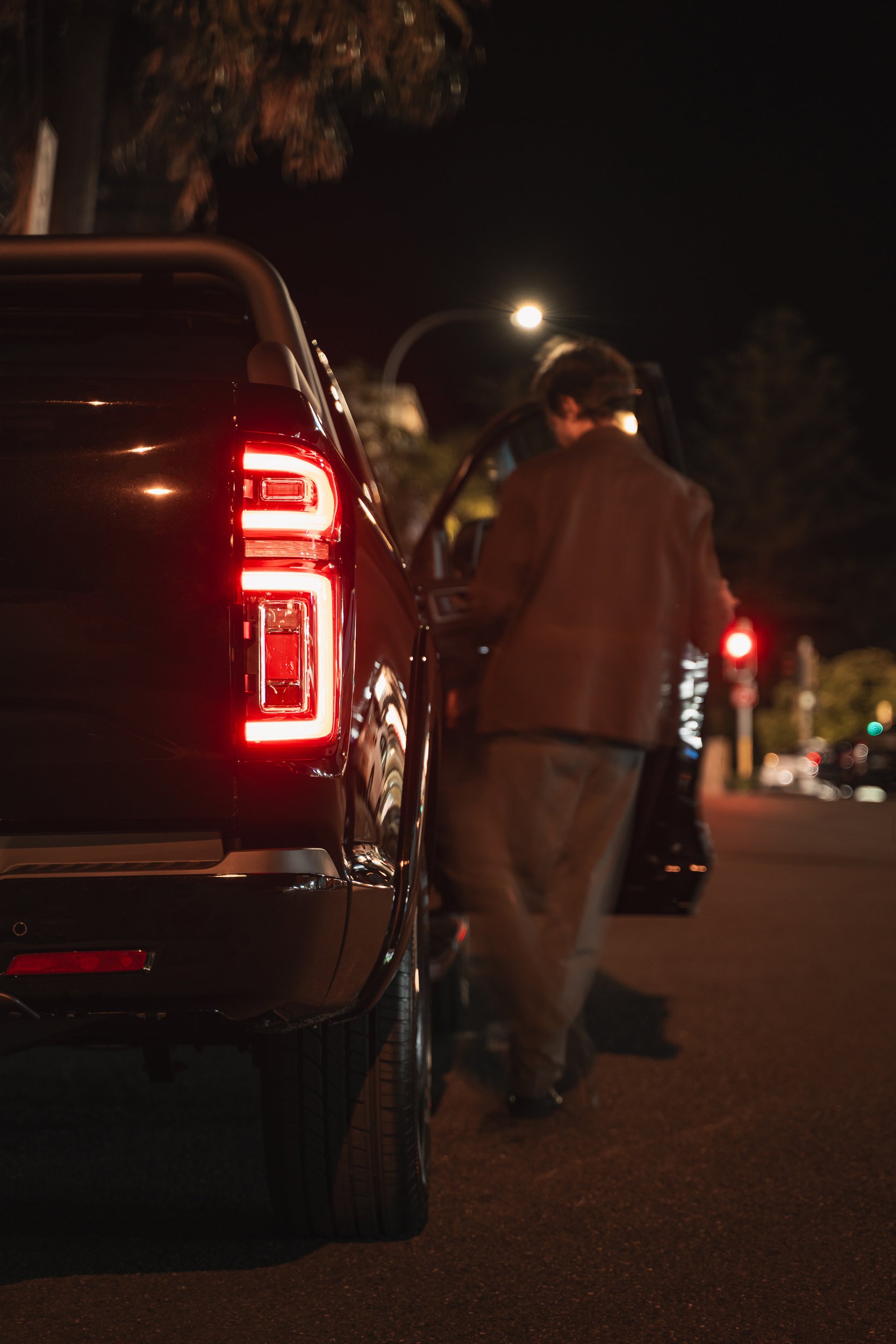 A person standing next to a black pickup truck at night, with the truck's taillights illuminated, streetlights, and blurred background.