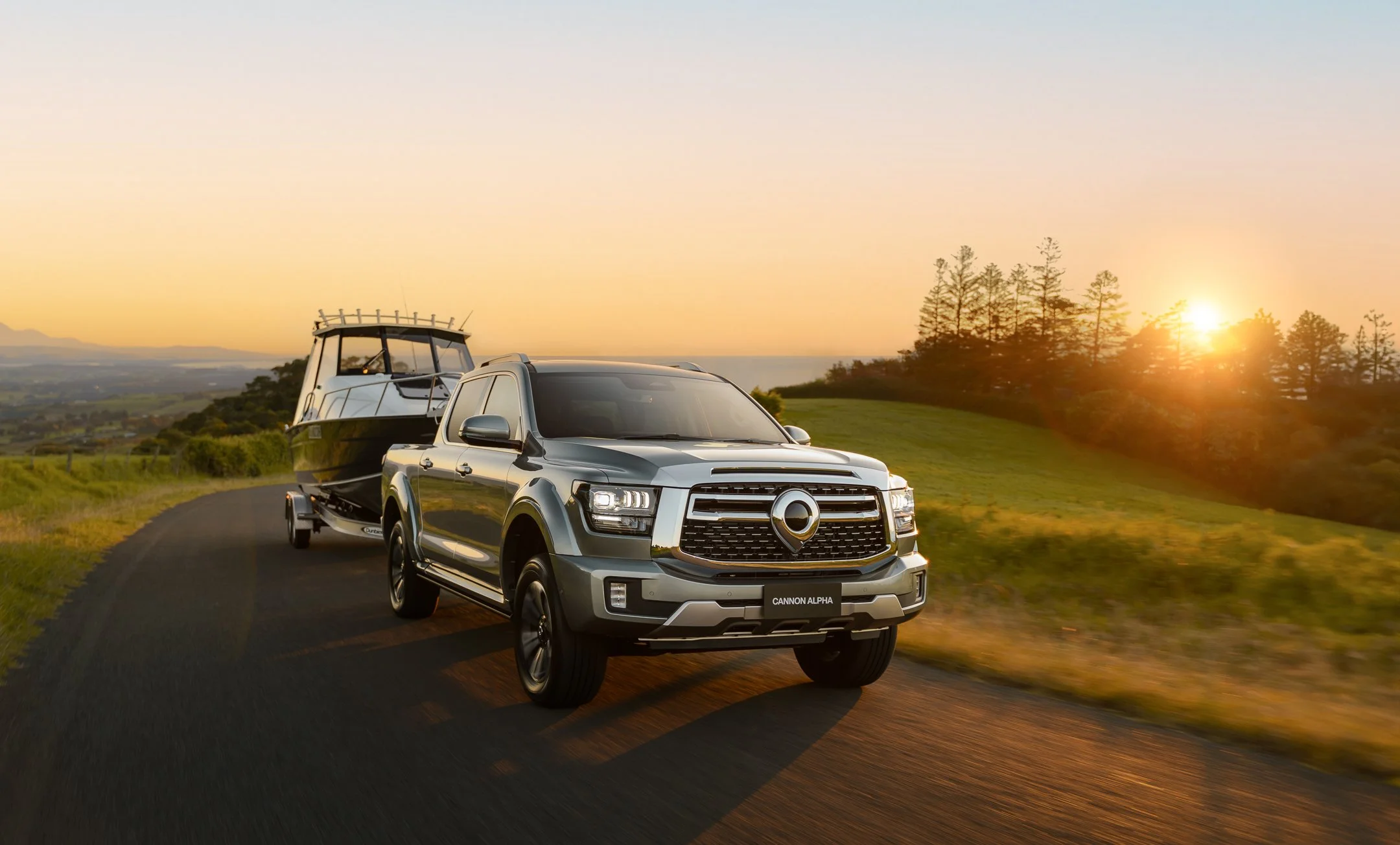 A silver pickup truck towing a boat on a trailer along a winding countryside road at sunset, with trees and distant mountains in the background.