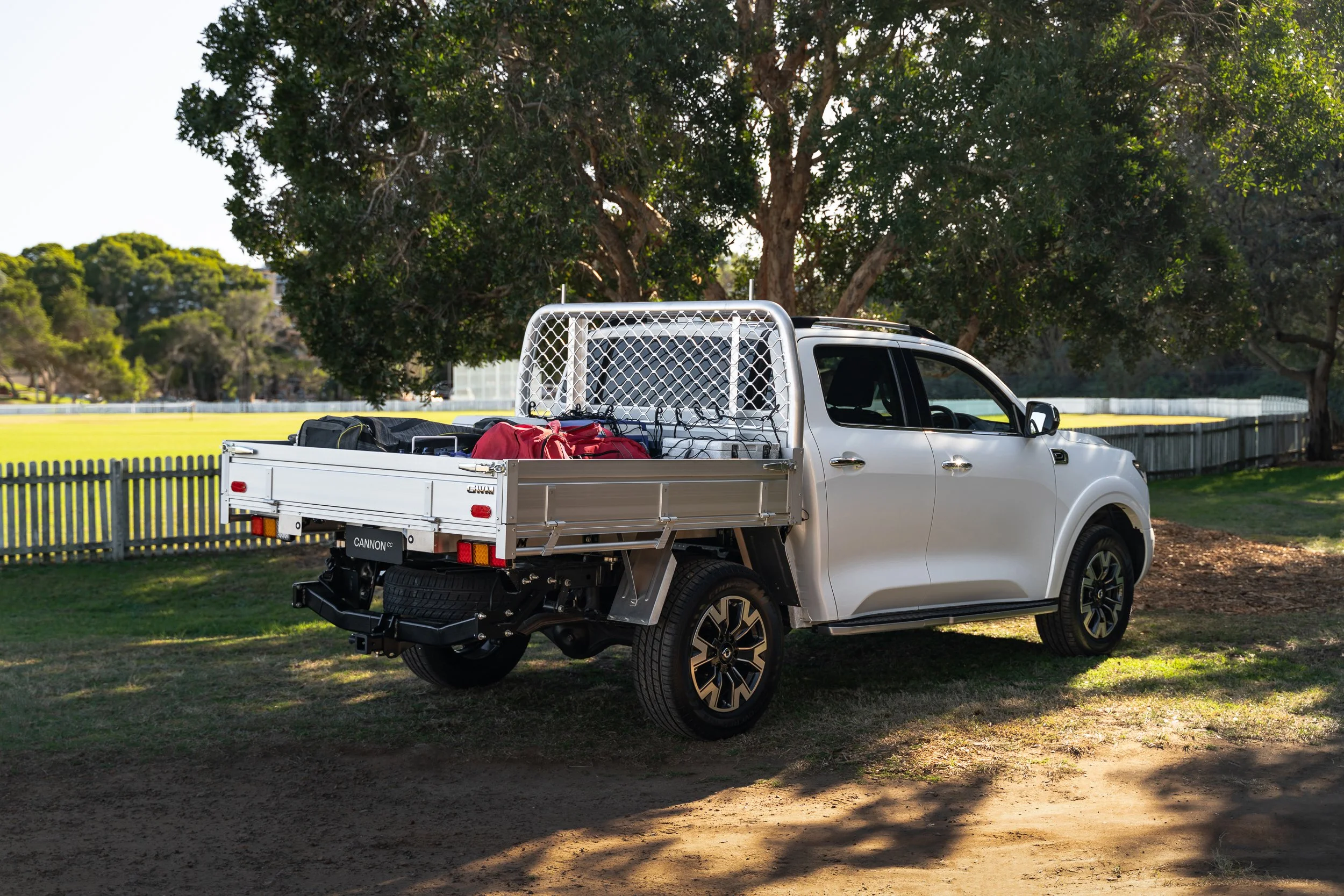 A white pickup truck parked on grass next to a fence and large trees, with a clear sky in the background, carrying bags and equipment in its open cargo bed.