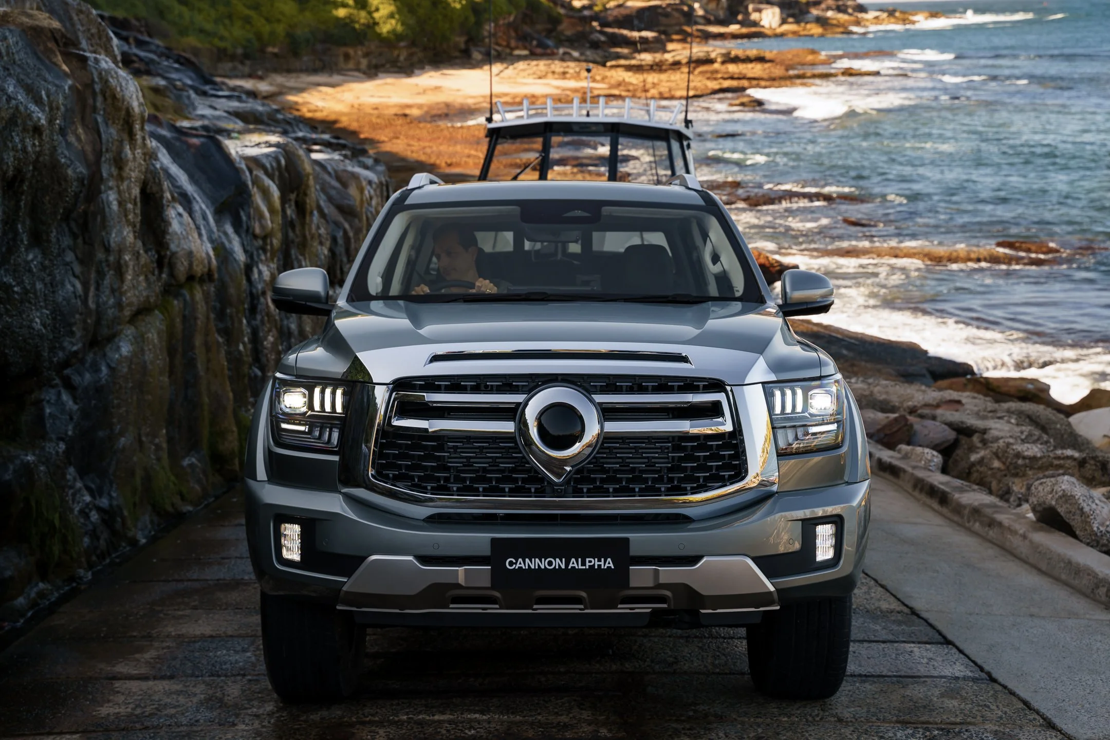 A silver Cadillac SUV parked along a rocky coastline with a waterfall on the left and ocean waves in the background, featuring a black license plate with white text that reads 'CANNON ALPHA.'