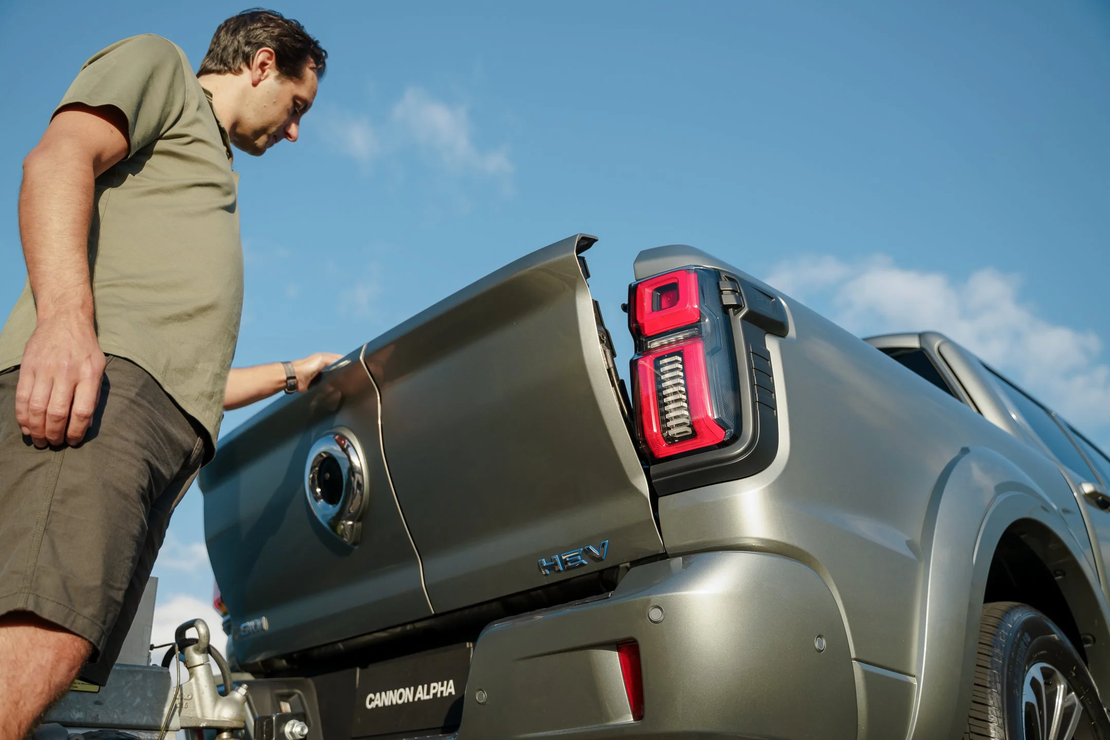A man standing beside a gray pickup truck with the tailgate open, looking into the truck bed against a clear blue sky.