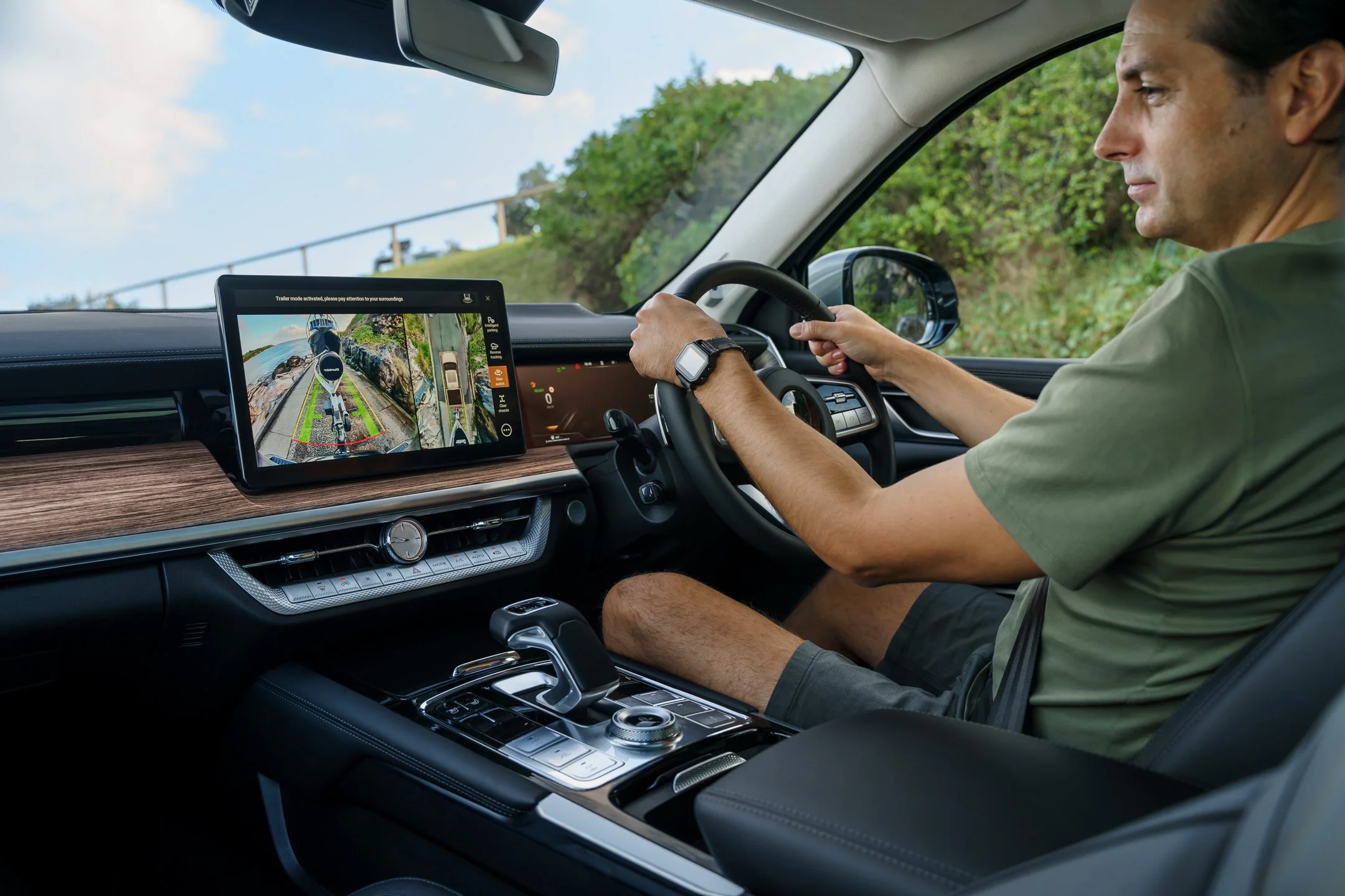 A man sitting in the driver's seat of a vehicle, operating the steering wheel with his left hand. The vehicle's dashboard features a digital display showing a rearview camera feed with parking guidelines, and a tablet mounted in the center with a nav