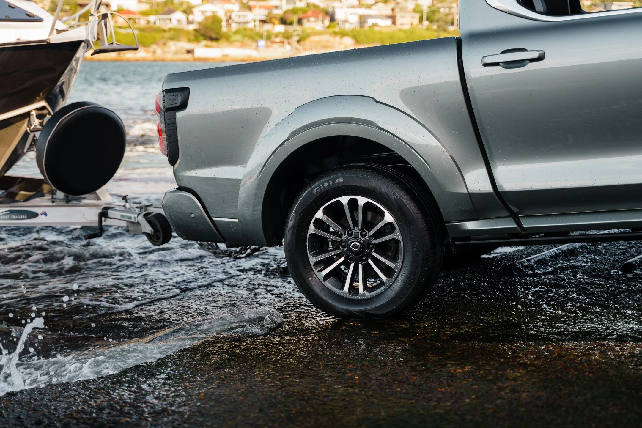 A silver pickup truck on a boat ramp, with water splashing around its tires, boat trailer attached, and a boat in the water nearby against a backdrop of a residential neighborhood on a hill.