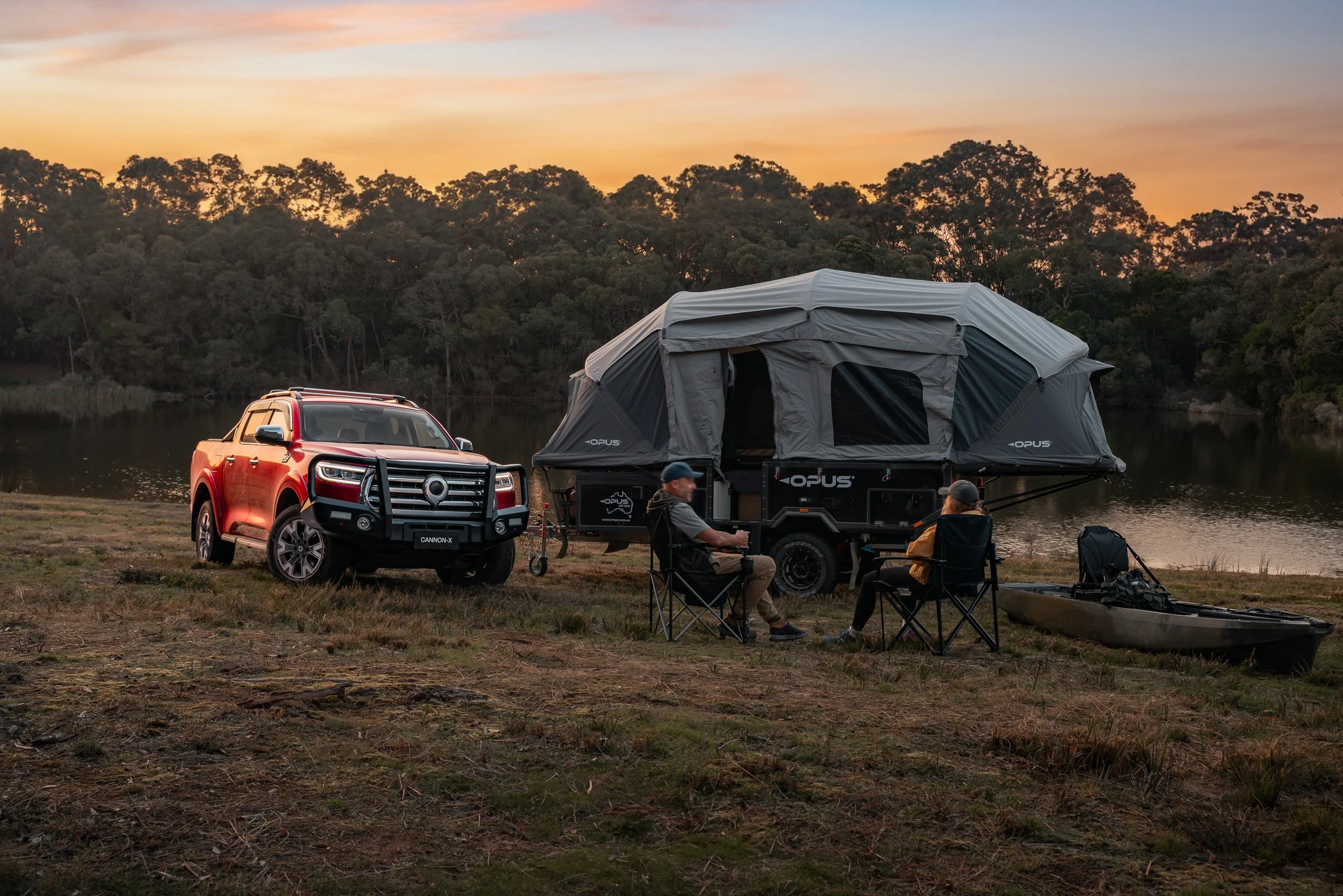 A red pickup truck parked on the grass near a lake at sunset. Next to the truck, there is a large camping tent on a trailer. Several people are sitting around in camping chairs, with a kayak on the ground nearby. The background features a lake and de