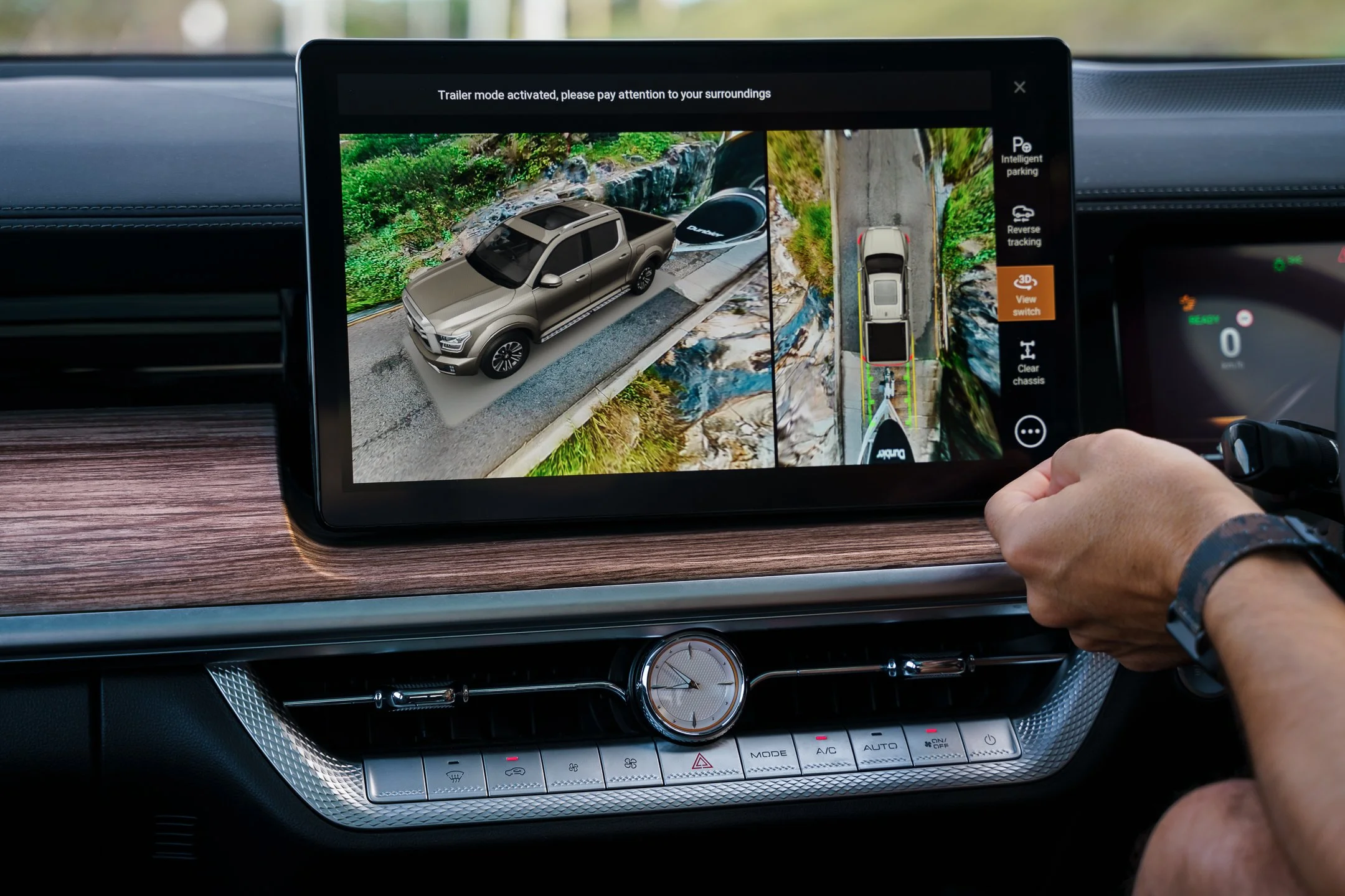 A car's dashboard and center console with an infotainment screen displaying a 3D view of a vehicle and its surroundings, including a river, rocks, and greenery.
