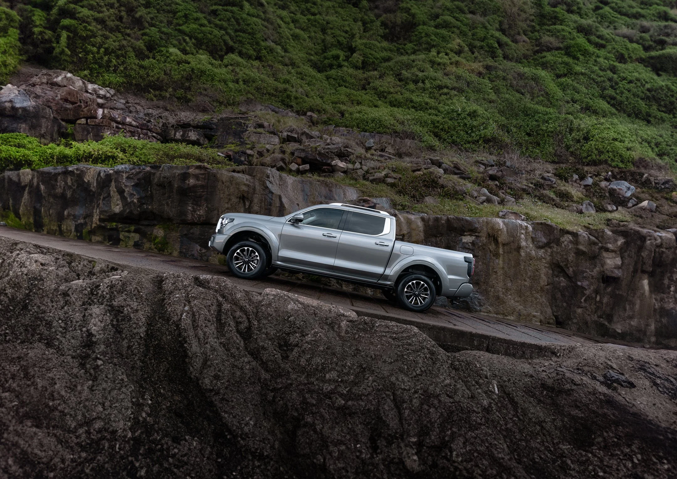 A silver pickup truck driving along a road next to a rocky hillside covered in green bushes and trees.