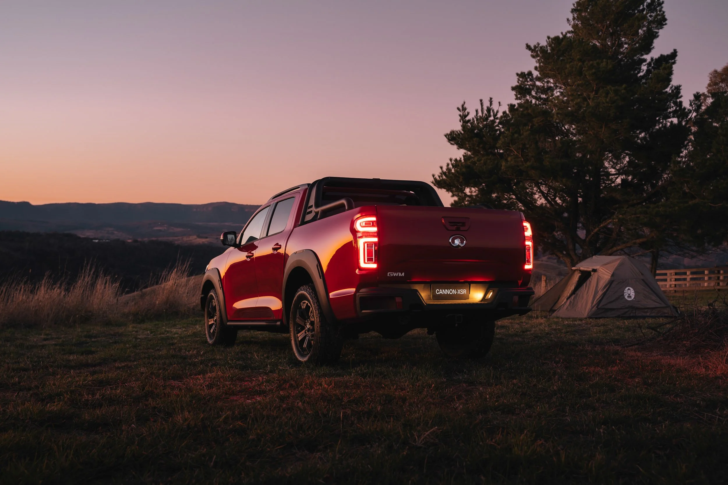 Red pickup truck parked on grass with a camping tent nearby, trees and a mountain range in the distance during sunset.